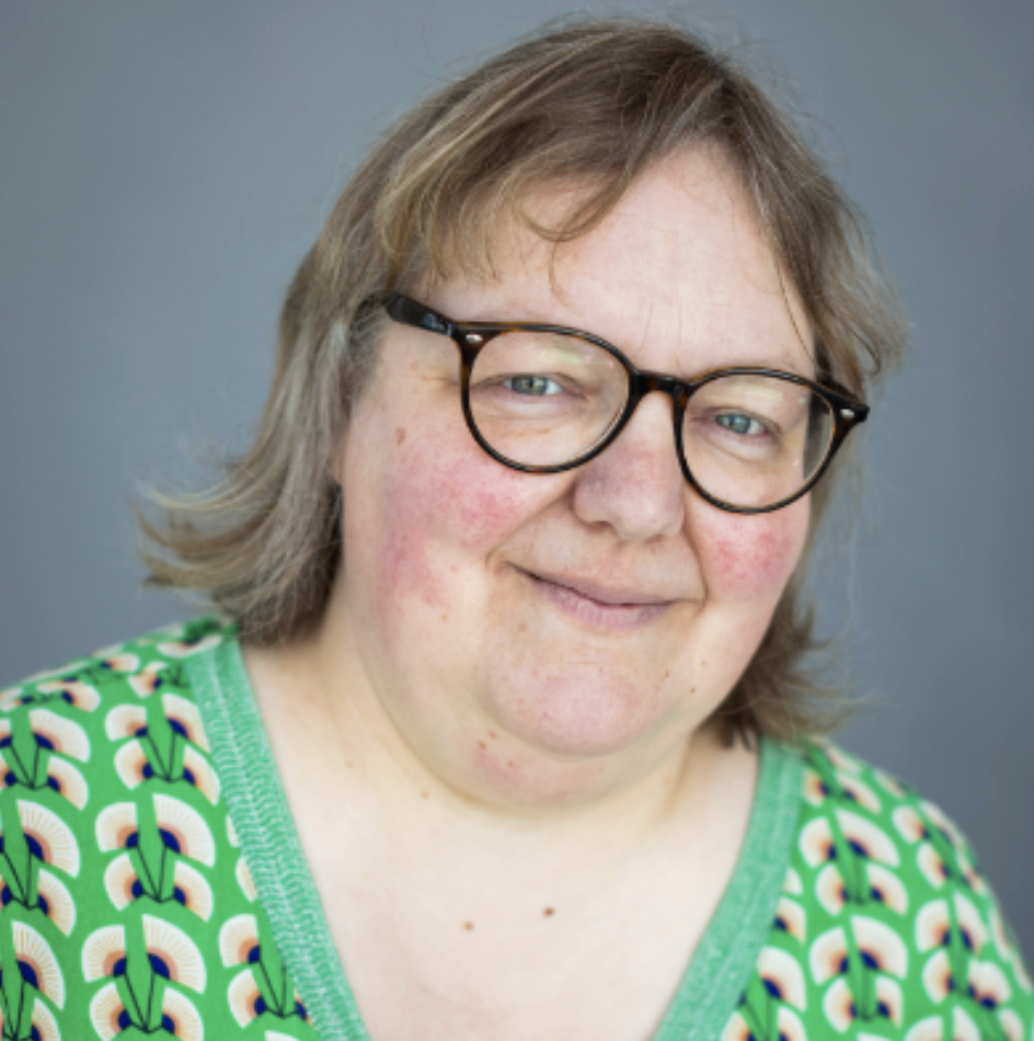 Portret van een persoon met bruin haar, brillen, en een groene shirt met patroon, lachend voor een grijze achtergrond.