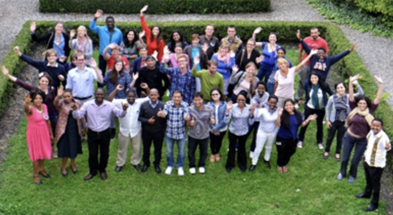 Een grote groep mensen die samen buiten in een tuin staan, vriendelijk lachen en zwaaien.