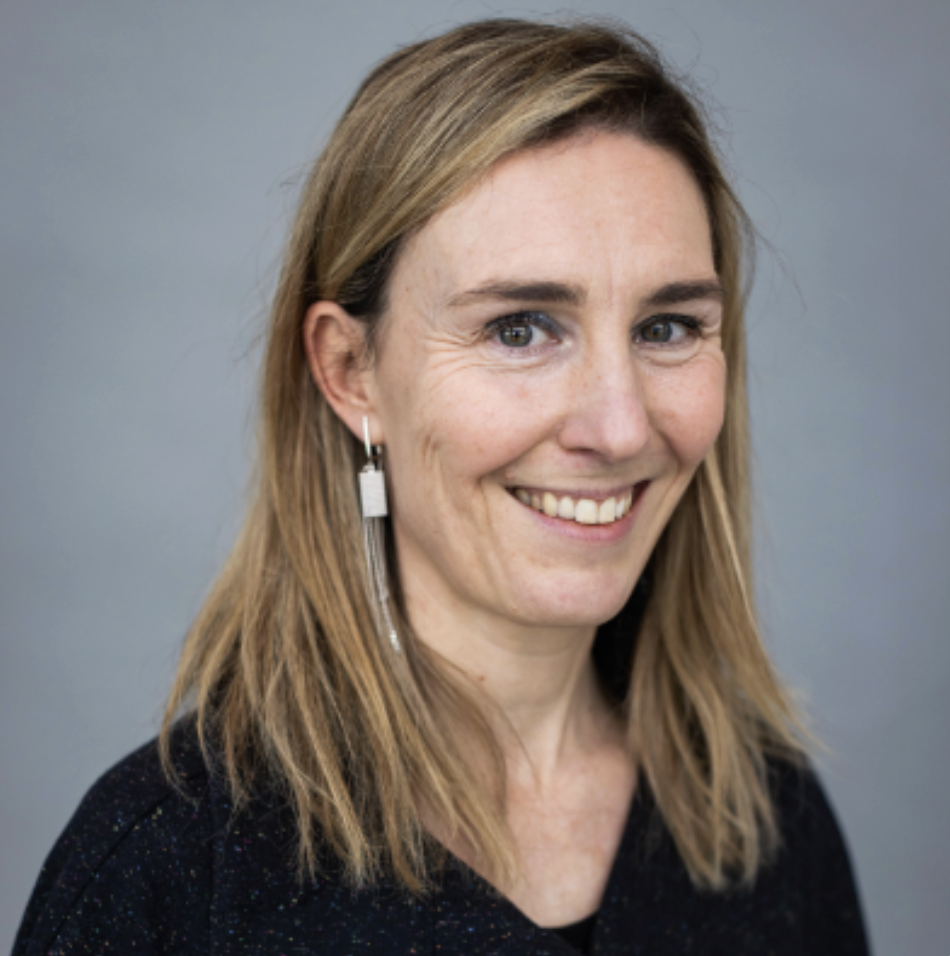 Portret van een lachende vrouw met schouderlange, lichtbruine haar, blauwe ogen, witte tanden, een zwart shirt en lange oorbellen, voor een grijze achtergrond.