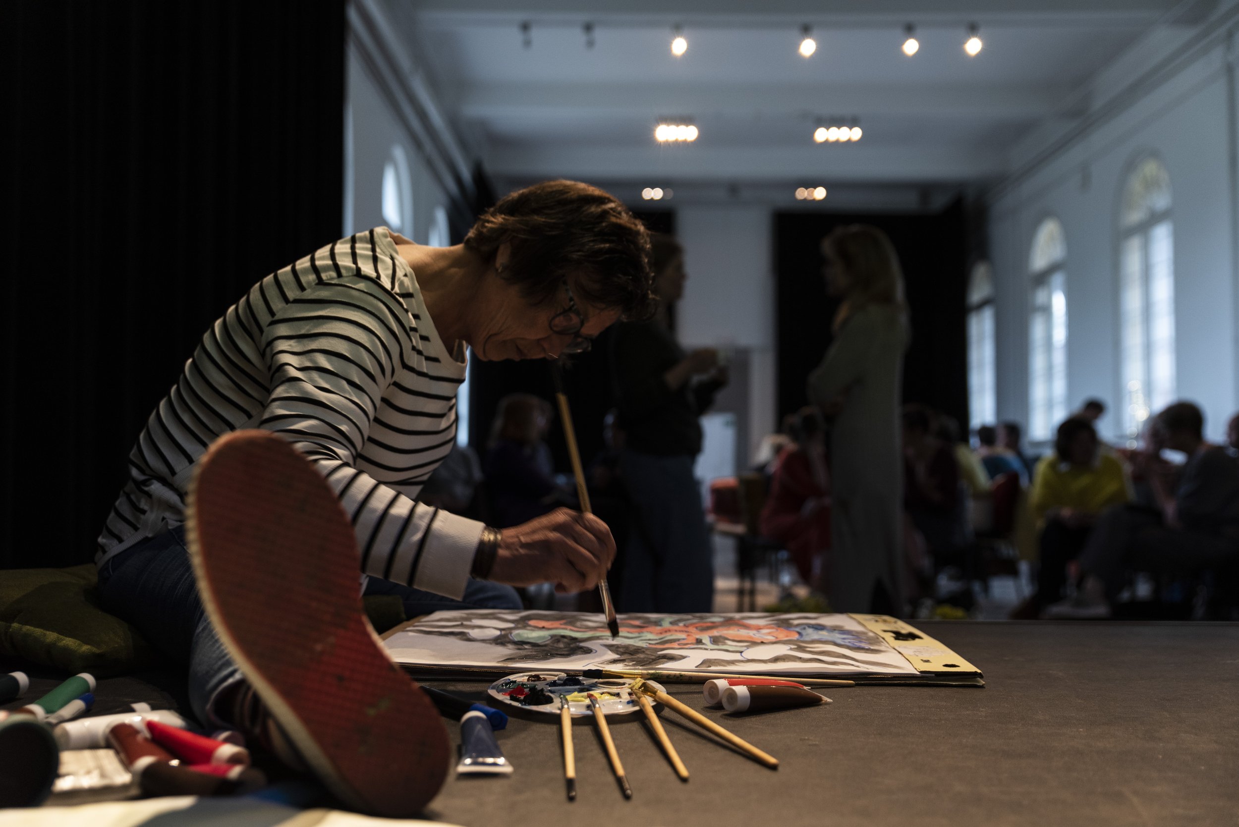 Vrouw die schildert in een grote ruimte met mensen op de achtergrond, kunstmaterialen op tafel