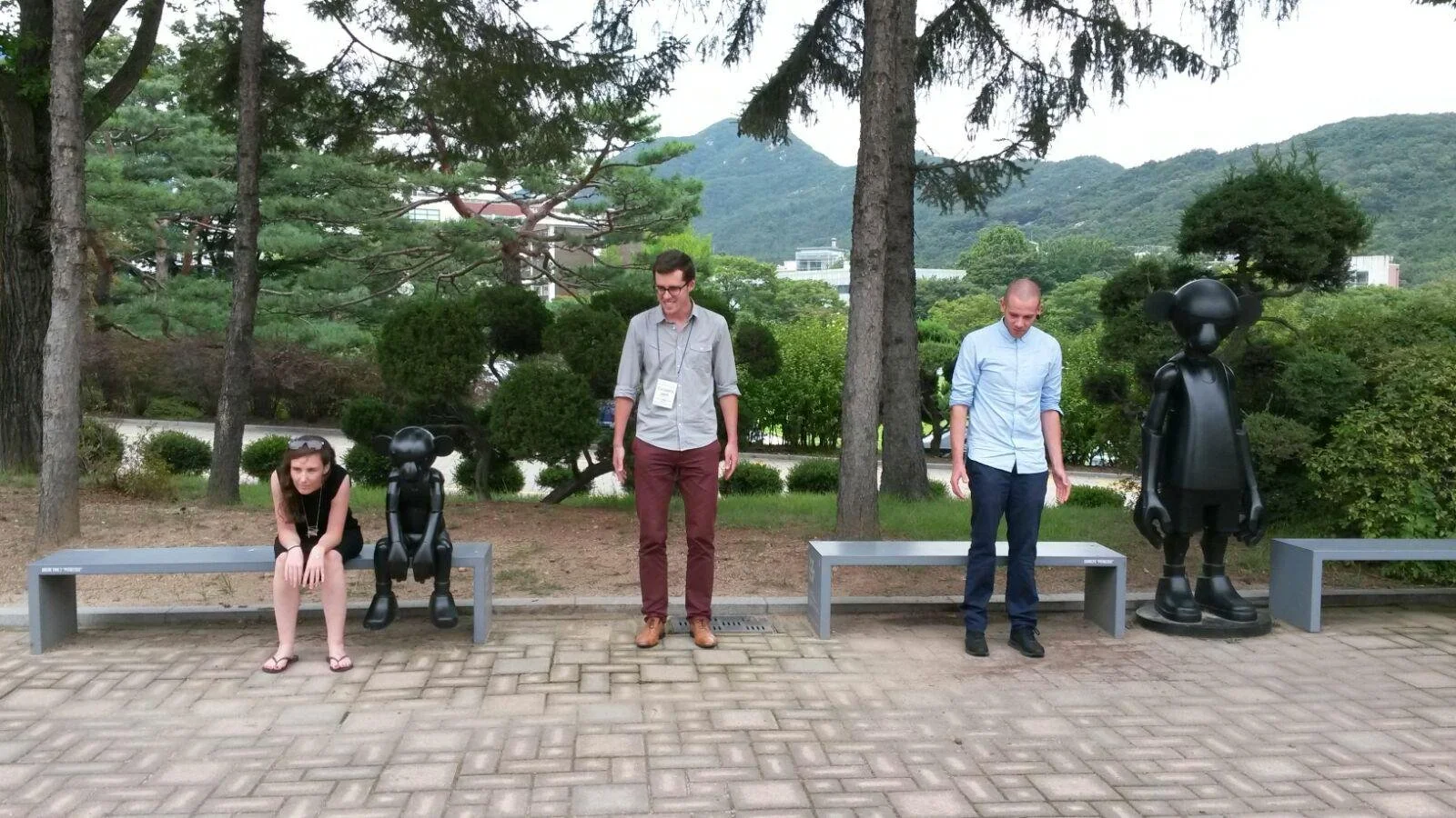 Twee mensen en twee beeldjes geïnspireerd door Mickey Mouse, zittend op banken in een park, met bomen en heuvels op de achtergrond.