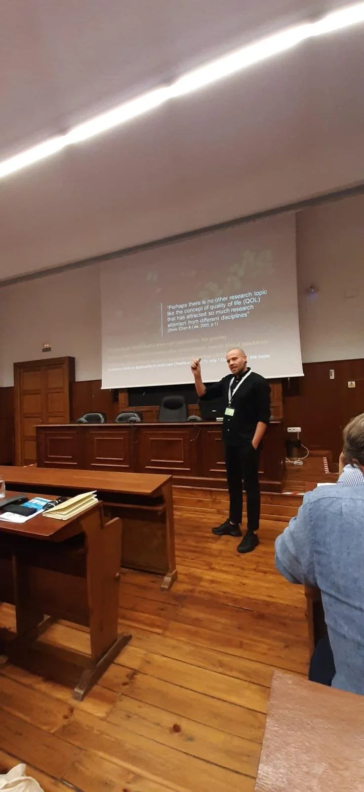 Een man geeft een presentatie in een houten kamertje met een groot projectiescherm, waar tekst wordt weergegeven, terwijl een publiek toekijkt.