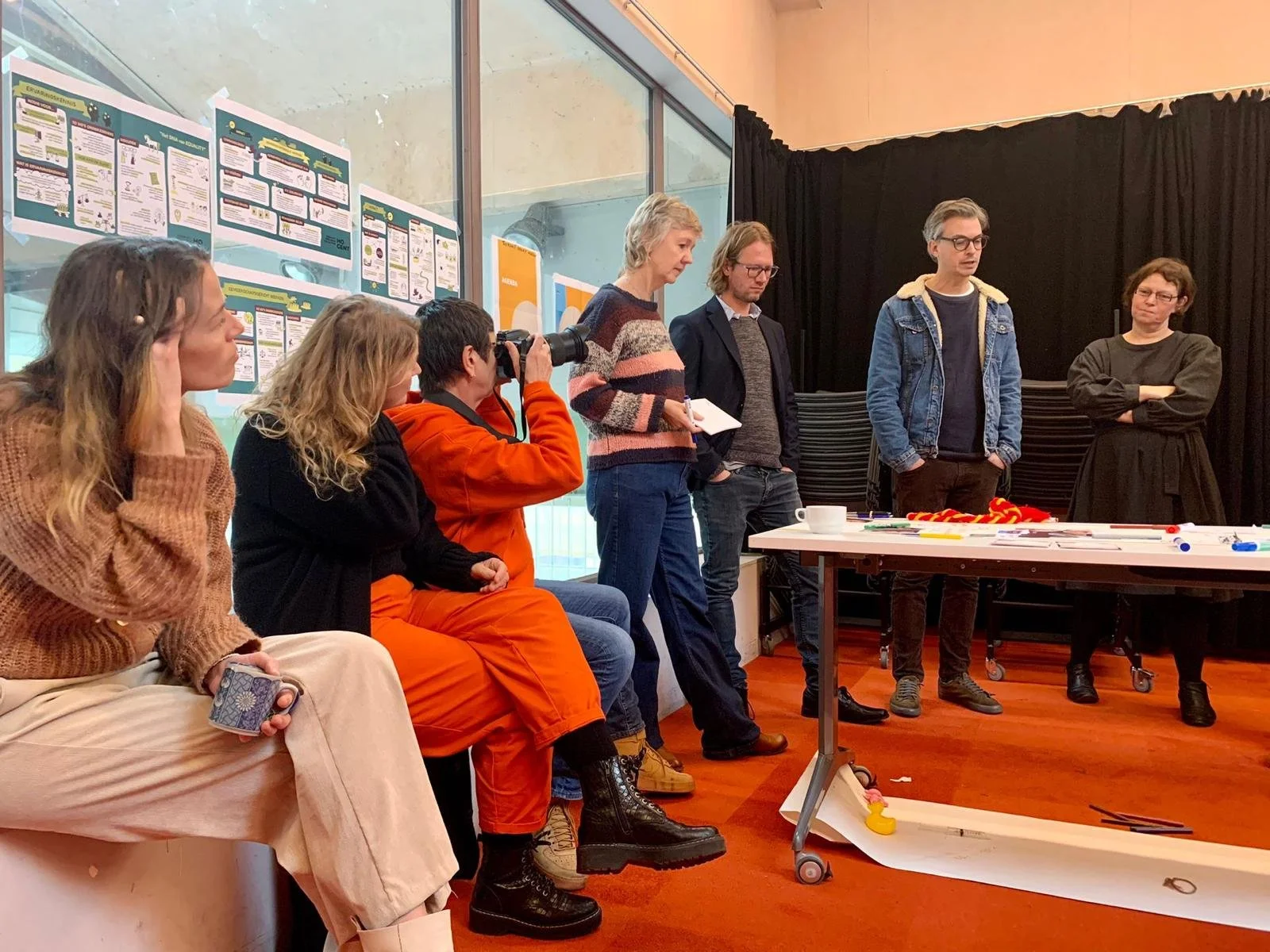 Vijf mensen staan en zitten in een kamer met een tafel met kleurrijke voorwerpen en een beker. Er hangen posters aan de muur.