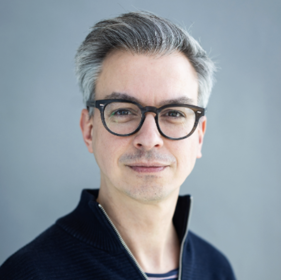 Portret van een man met grijs haar, zwarte bril en een blauwe sweater met rits, lachend voor een grijze achtergrond.