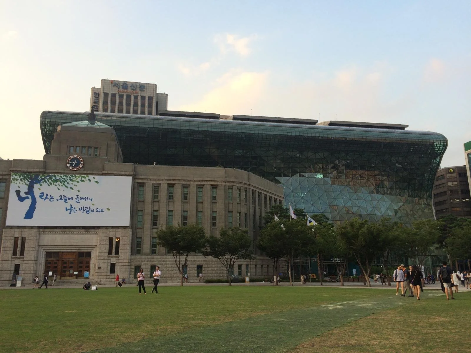 Een modern gebouw met een glazen gevel en een oud uitziend gedeelte met een klok. Mensen lopen en zitten op het gras in de voorgrond.