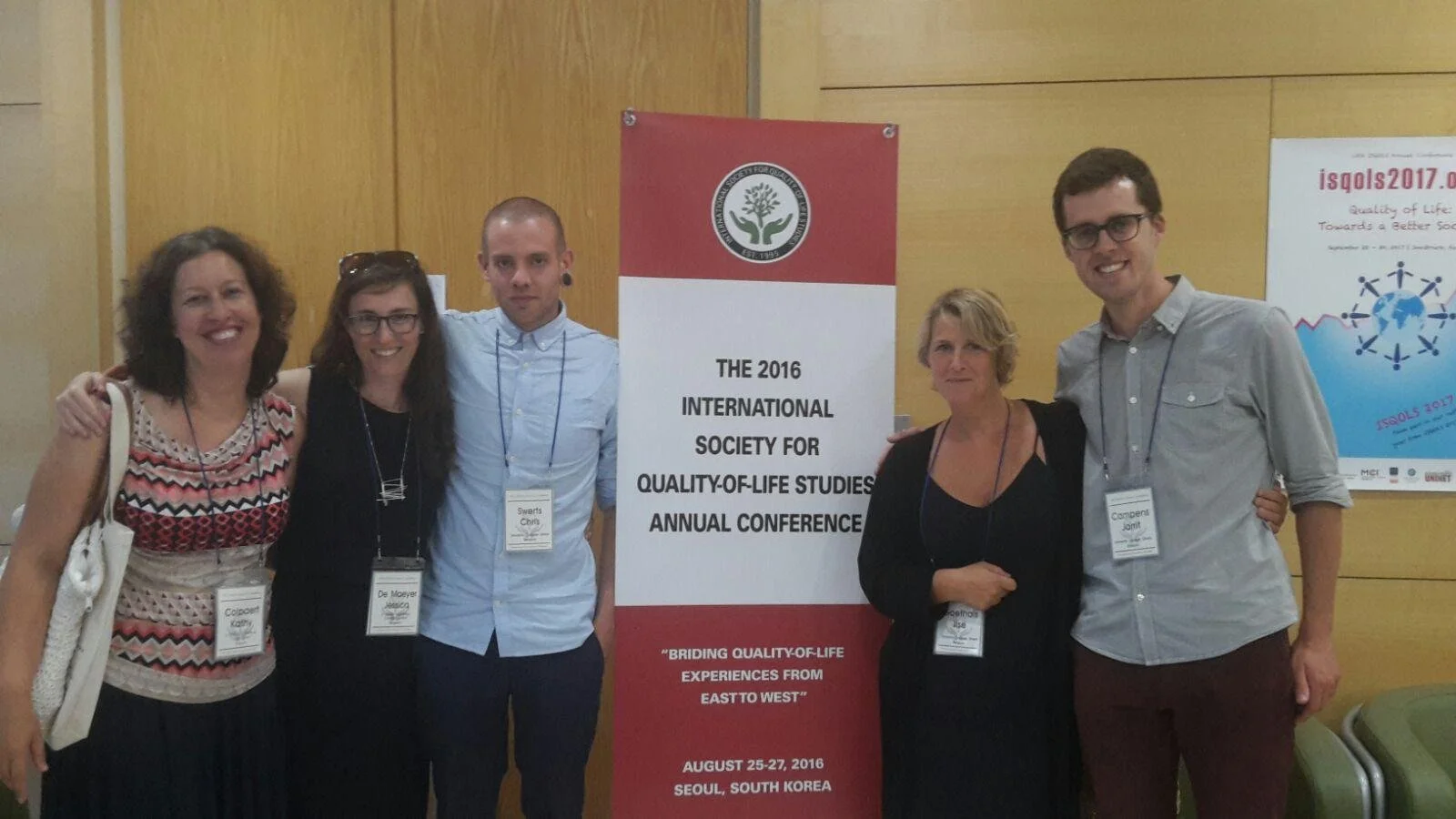 Vijf mensen staan samen bij een congres in Seoel, Zuid-Korea, met een banner op de achtergrond die verwijst naar de International Society for Quality-of-Life Studies in 2016.