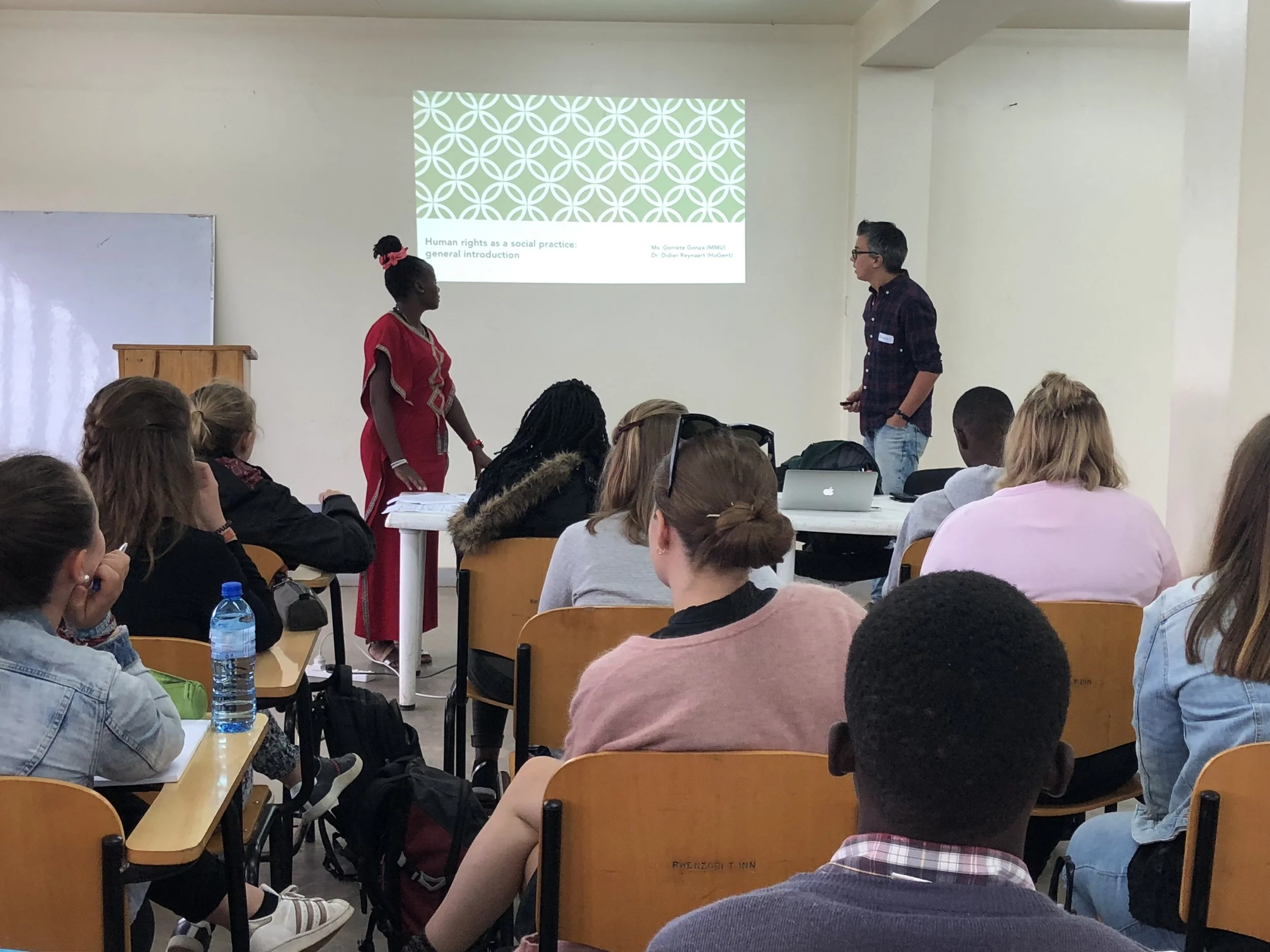Een klaslokaal met studenten die naar een presentatie kijken; een man en een vrouw staan voor de klas voor een projectiescherm.