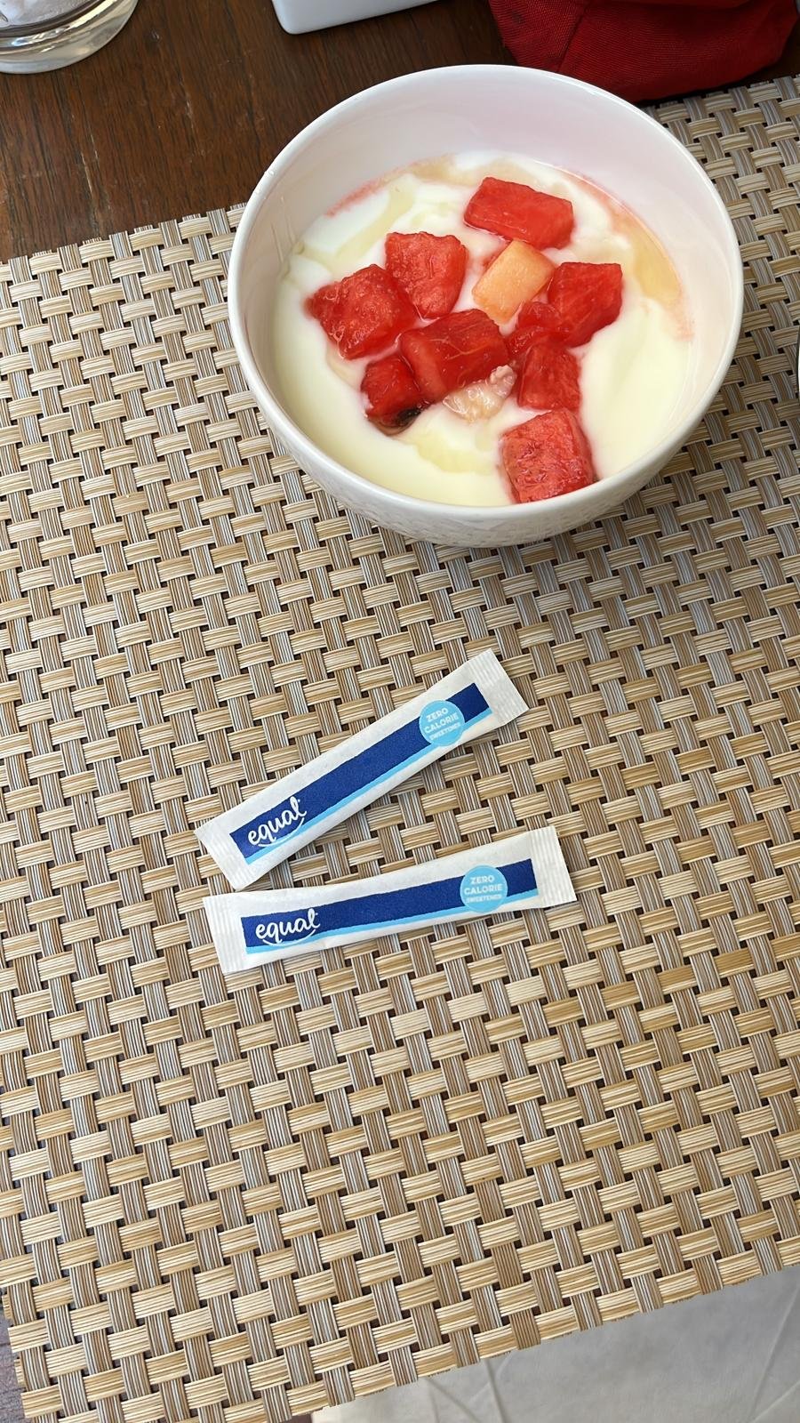 Een kom gevuld met yoghurt en stukjes watermeloen op een tafel met een rieten placemat en twee Equal suikerzakjes eronder.
