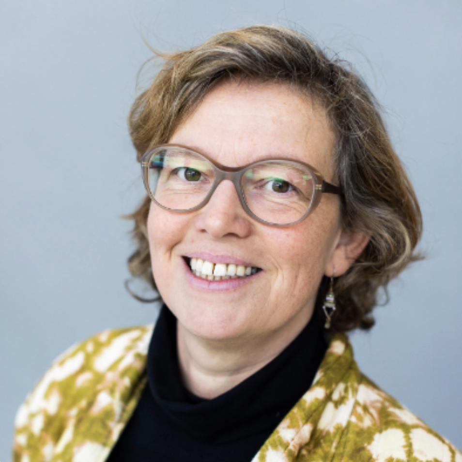 Portret van een lachende vrouw met bruin haar, glazen en een gebloemde jas, voor een grijze achtergrond.