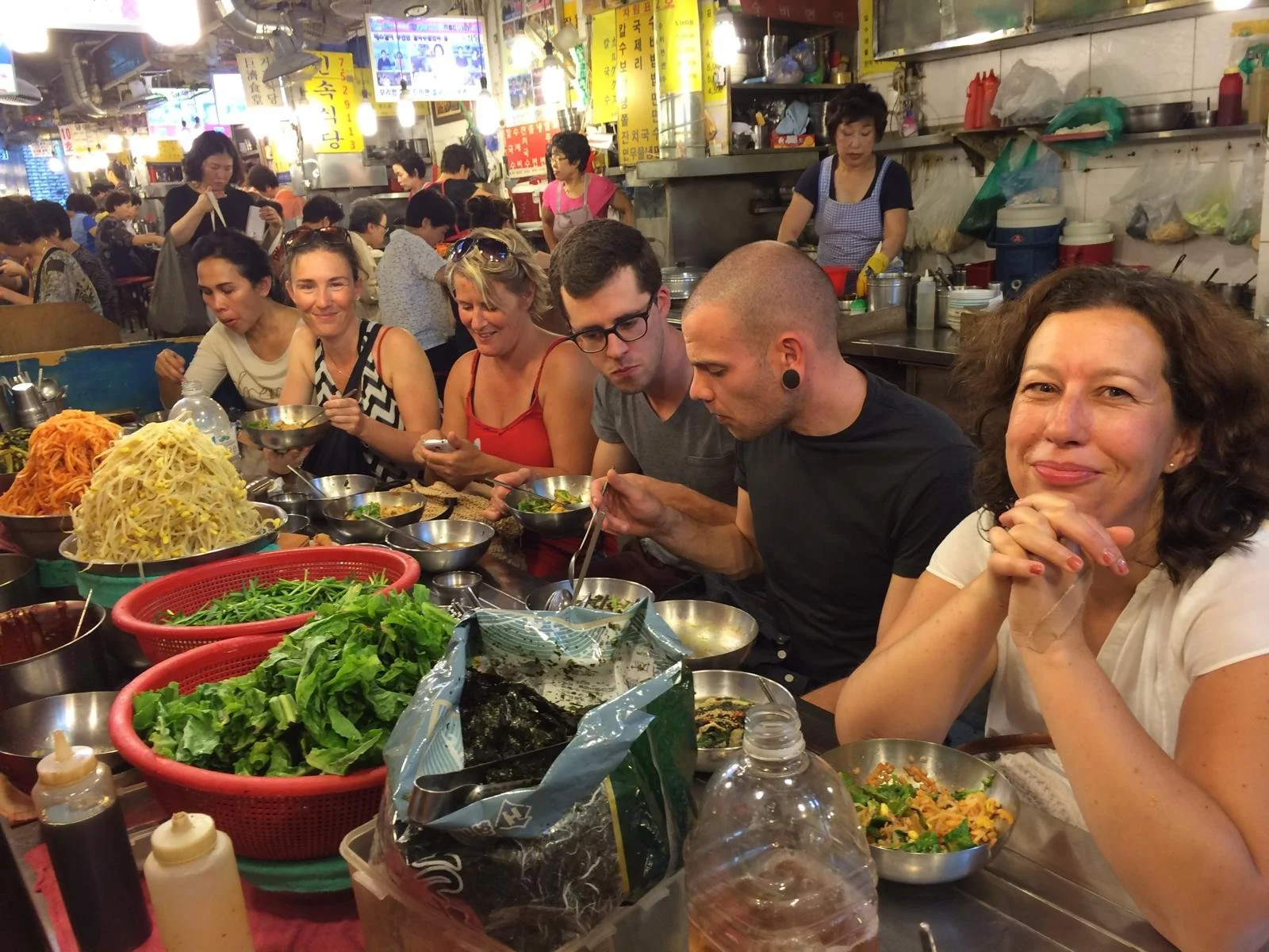 Vijf mensen zitten aan een tafel in een druk restaurant, met verschillende gerechten en groenten op tafel, terwijl ze genieten van hun maaltijd.