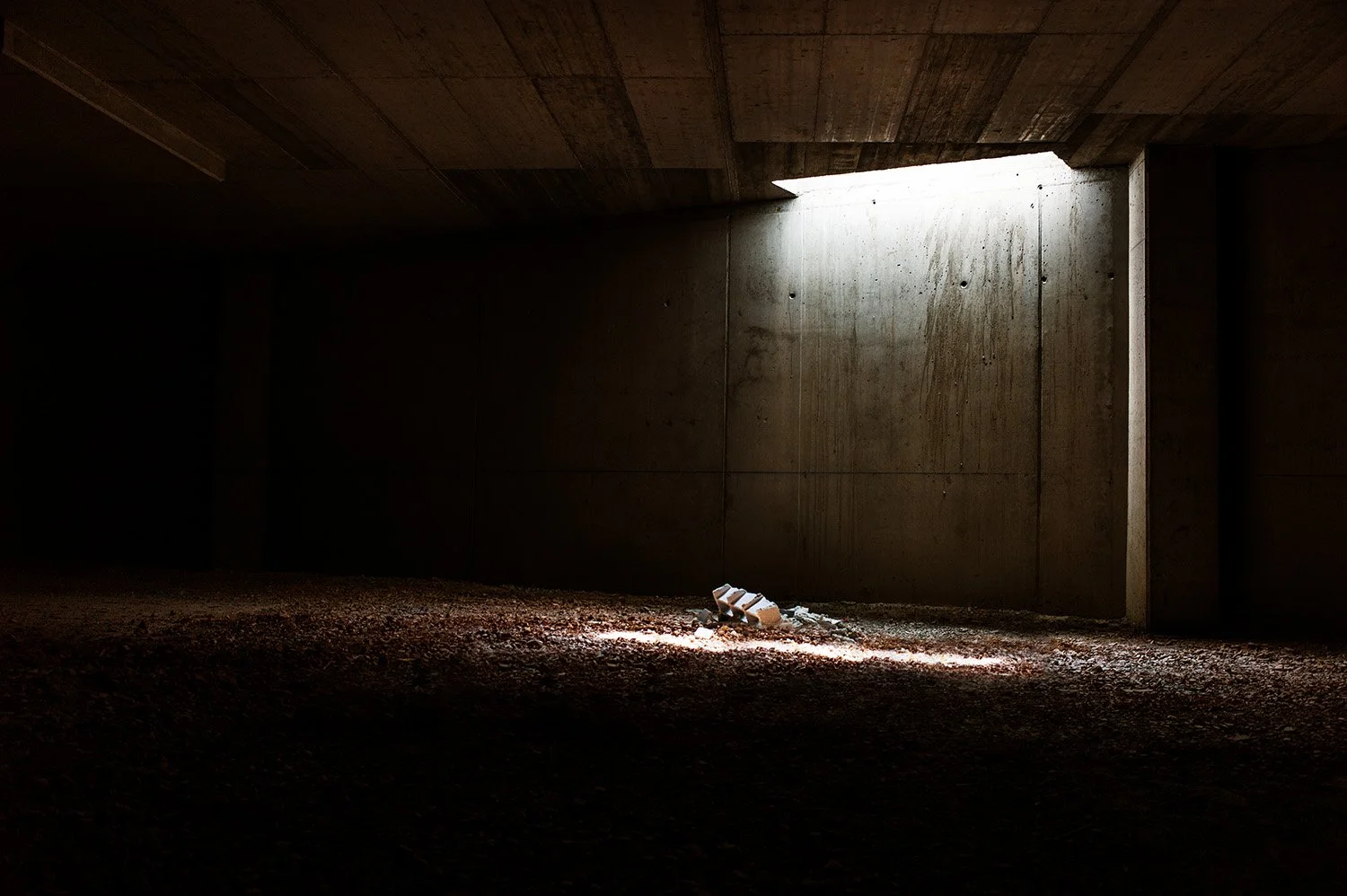 A dark, empty space with a concrete wall and ceiling, illuminated by a narrow beam of natural light from an opening at the top right, with some debris on the floor near the light
