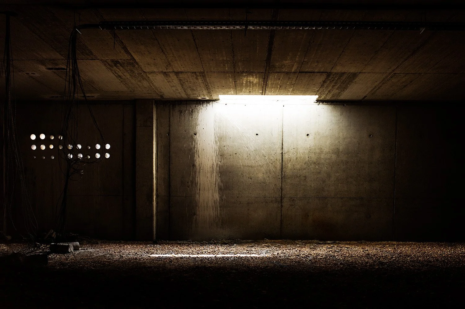 An underground parking garage with a concrete wall illuminated by natural light from an opening in the ceiling, with electrical wires hanging on the left side.