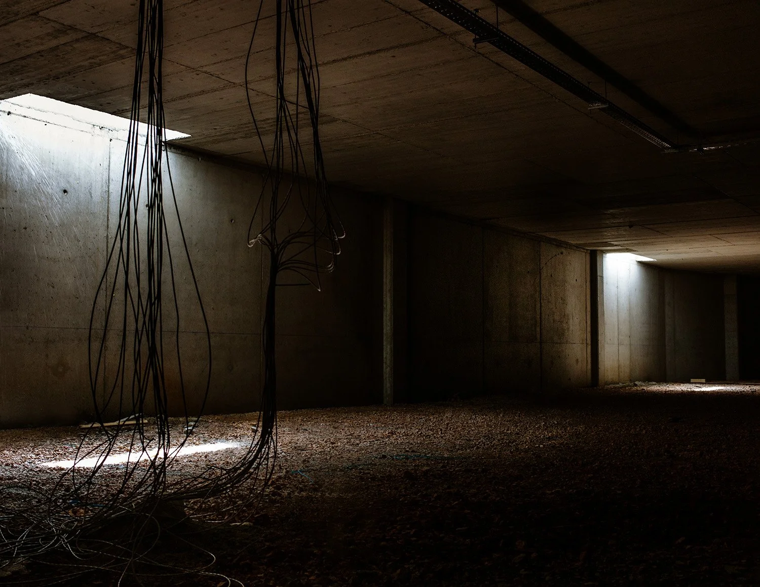 Interior of an unfinished concrete building with exposed wires hanging from the ceiling and small openings allowing natural light to come in.