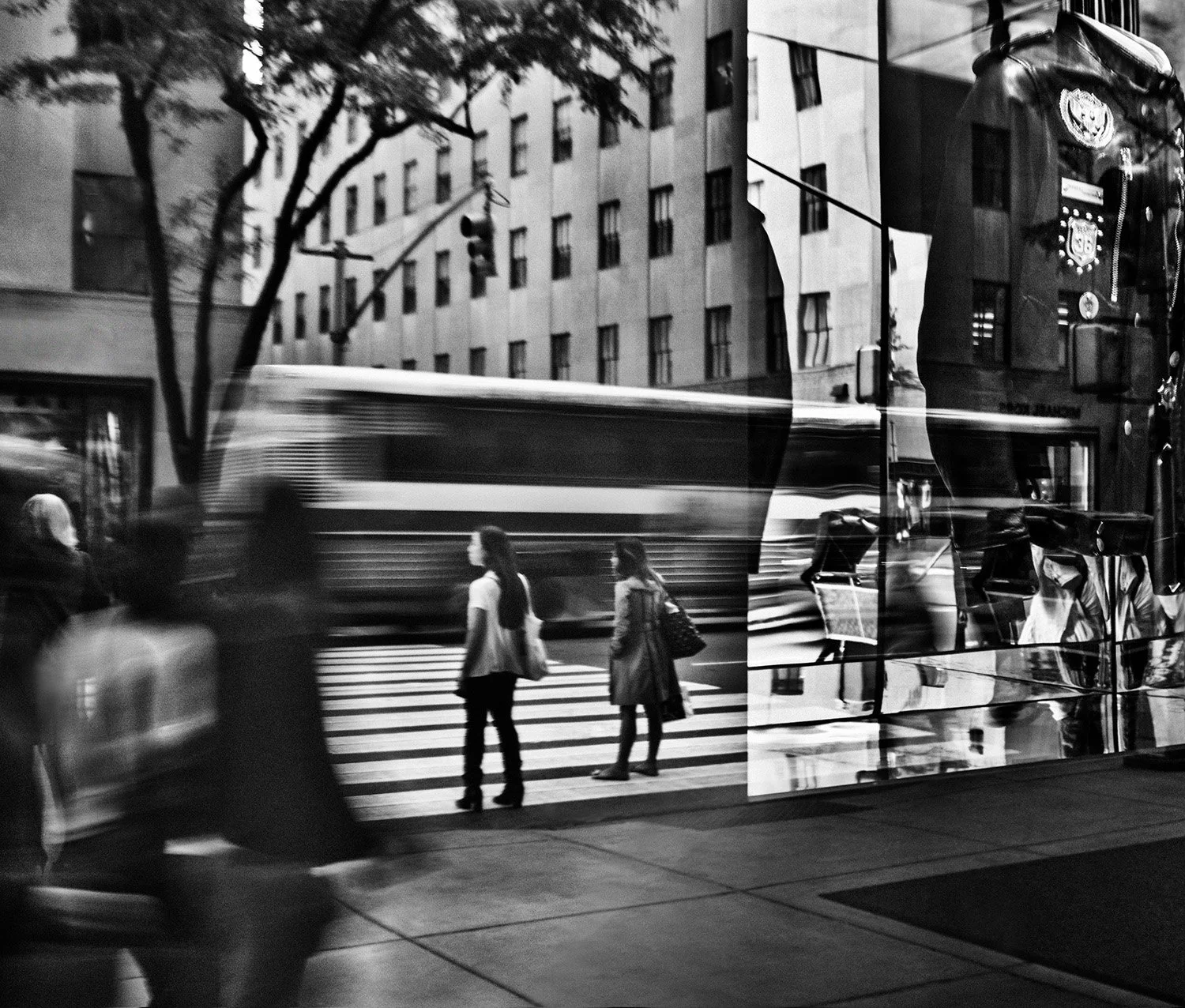 Black and white street scene reflections showing pedestrians waiting at a crosswalk, a moving bus in the background, a tall building, and trees, seen through a reflective surface on a storefront.