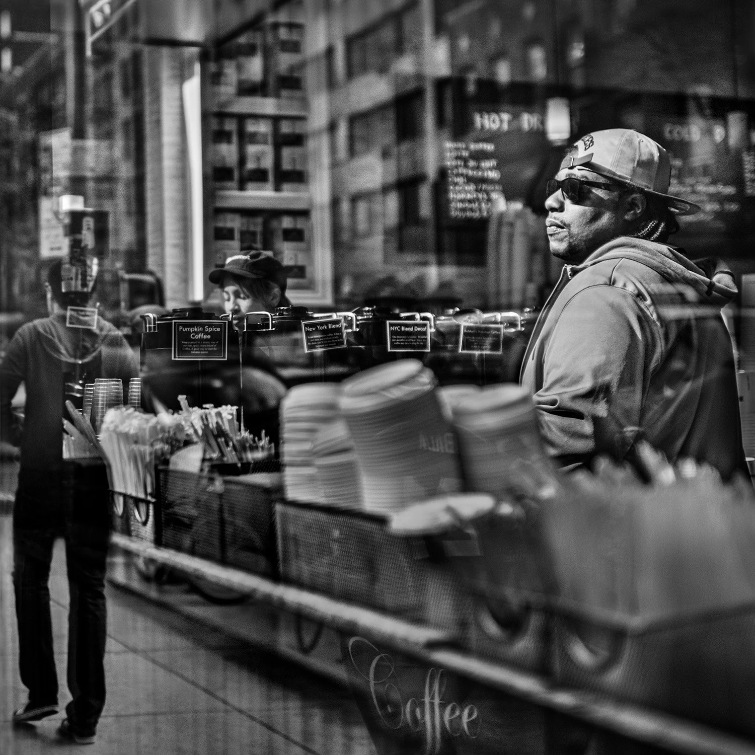 Black and white photo of a man standing outside a coffee shop, seen through the shop window. The man is wearing sunglasses, a cap backwards, and a hoodie. Inside the shop, there are signs on the counter displaying different coffee flavors.