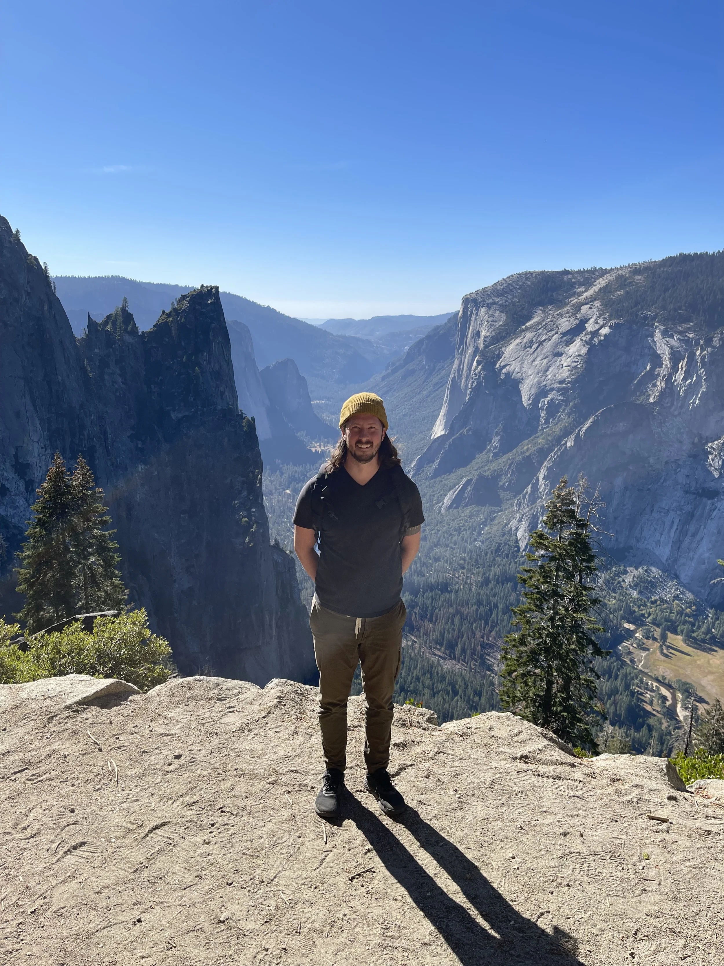A man standing on a rocky ledge with a backpack, smiling, in a mountainous landscape with tall cliffs, pine trees, and a clear blue sky.