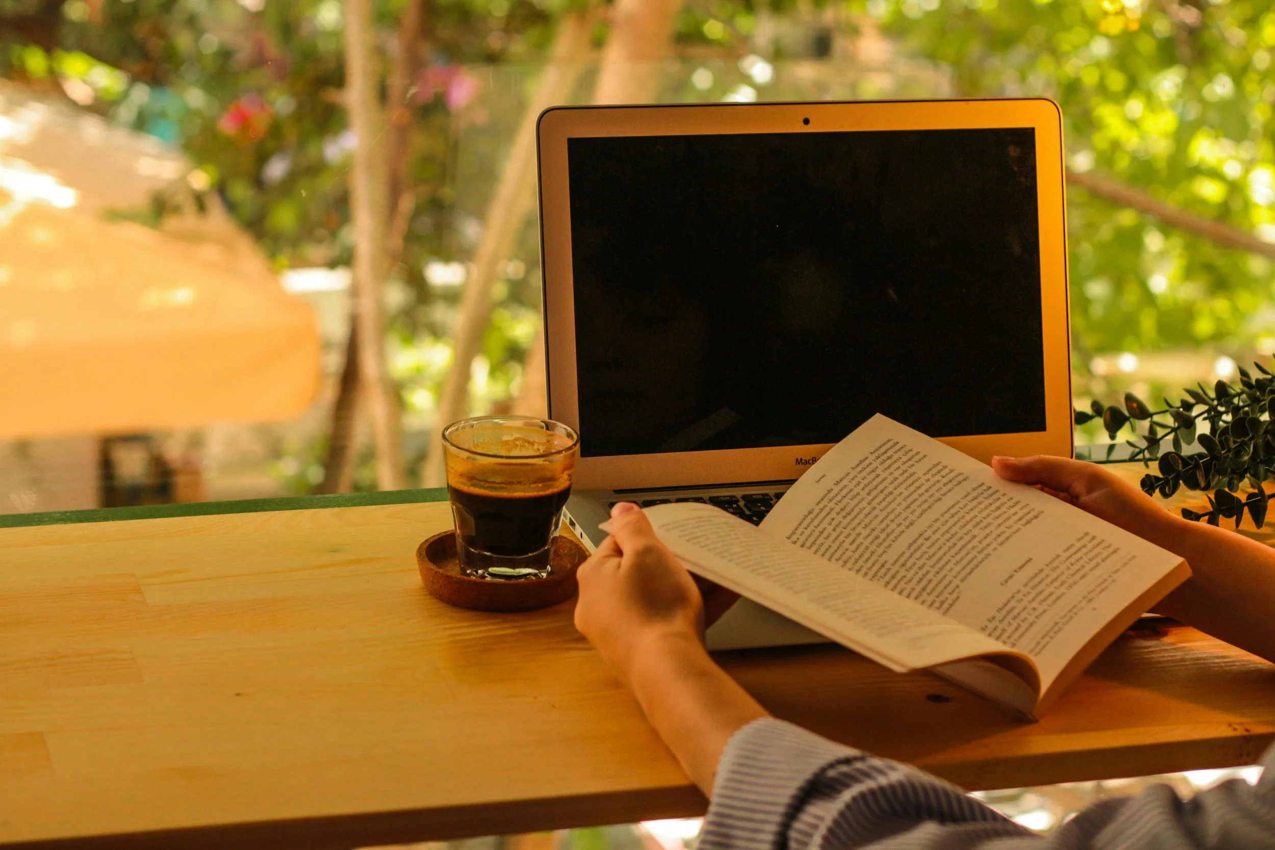 Person reading a book at a wooden table with a laptop and a glass of coffee or tea, outdoors with green foliage in the background.