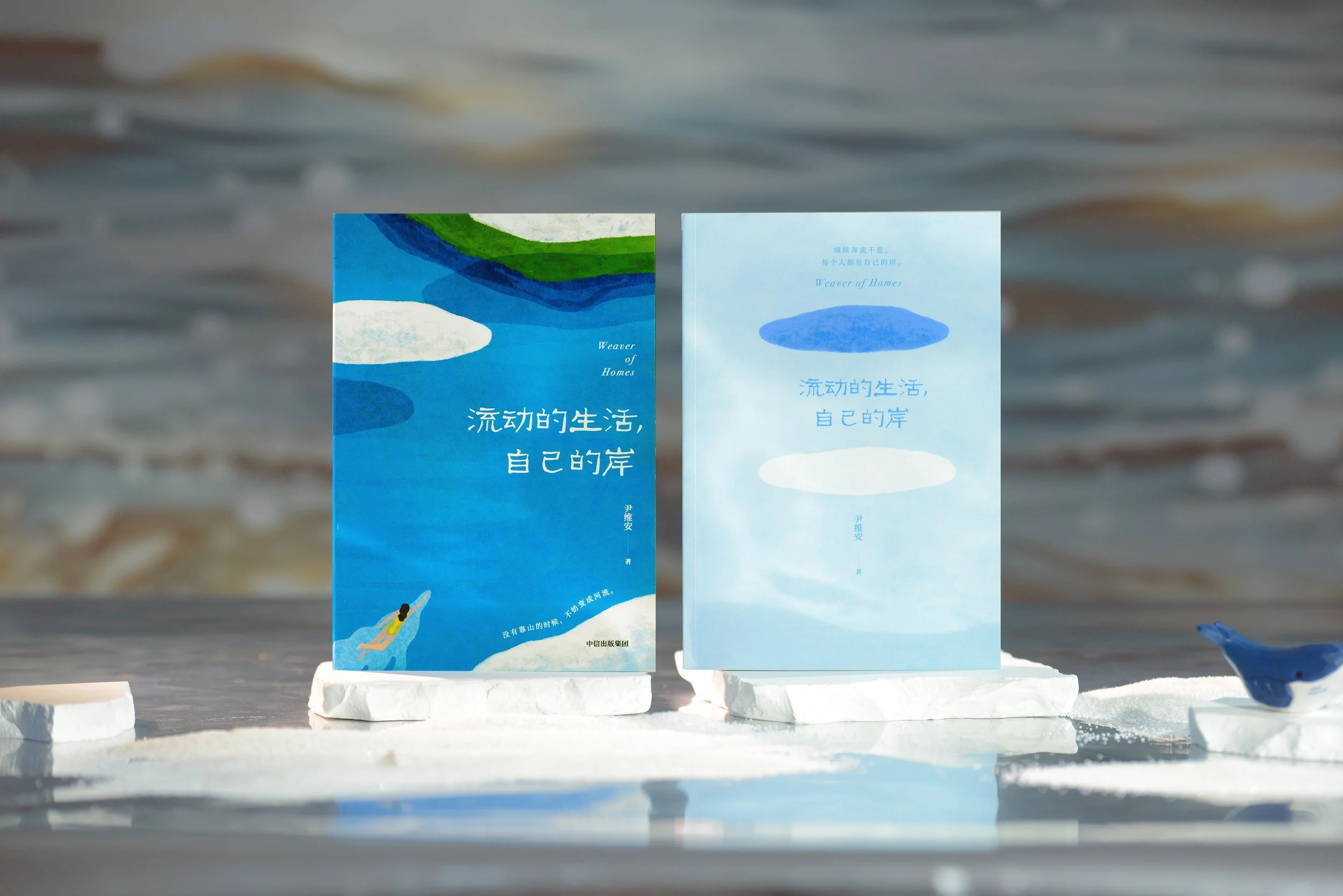 Two books titled "Weaver of Homes" with Chinese titles and cover art featuring watercolor clouds and a person swimming, displayed on white platforms, against a blurred ocean background.