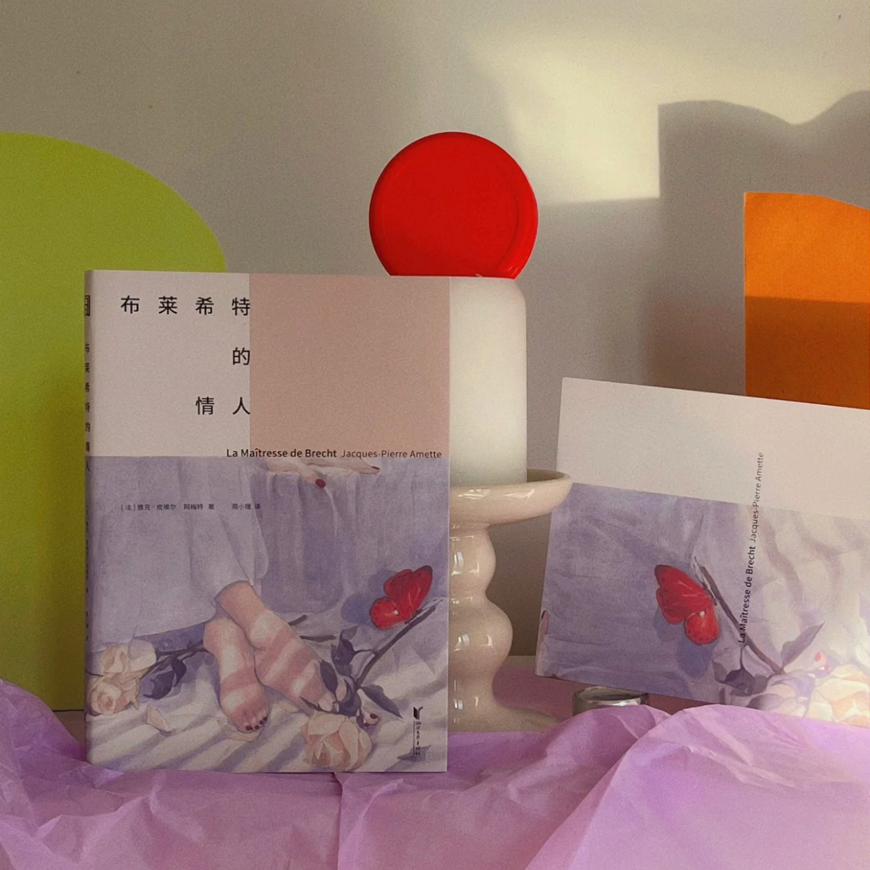 Books with cover art of a person in socks and a butterfly, positioned next to a lamp with a red circular top, set on pink tissue paper, with colorful abstract shapes in the background.