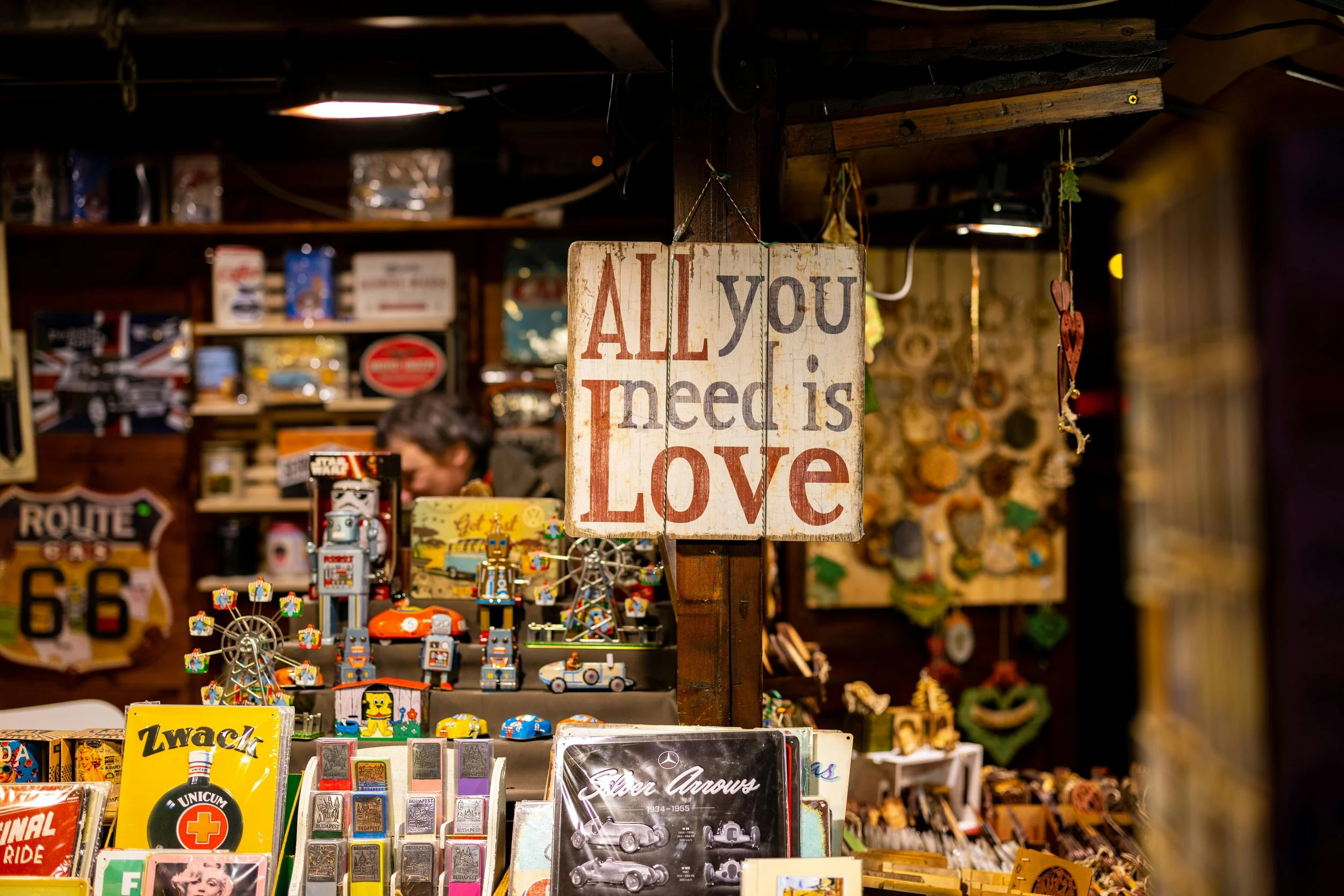 A vintage wooden sign reading 'All you need is Love' hanging in a shop filled with various toys, metal figurines, and decorative items, with a person blurred in the background.