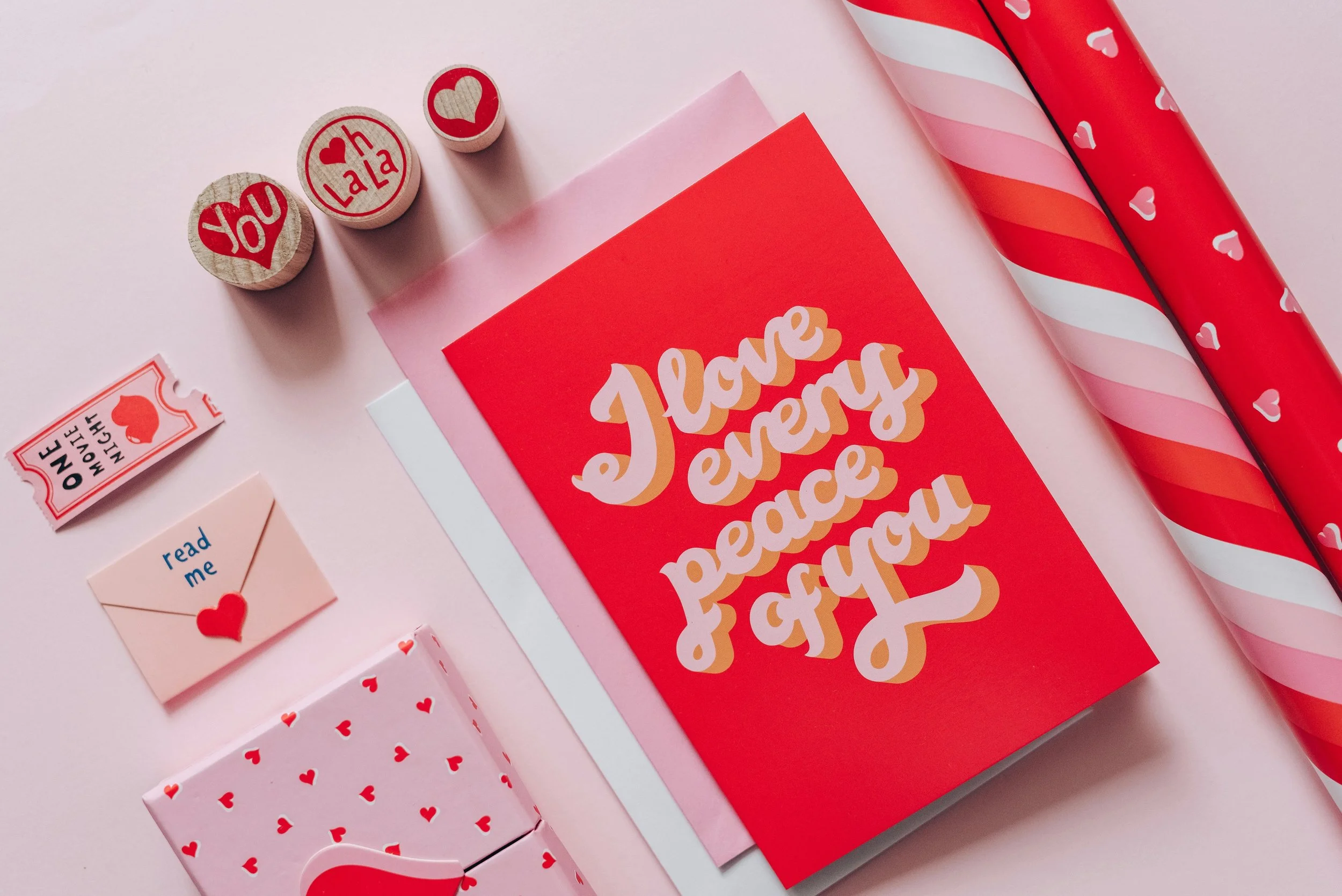 Valentine's Day gift setup with a red card reading "I love everyone & you," surrounded by love-themed decorations, including small stamps with hearts and words like "You" and "La La," a pink envelope with a red heart seal, and pink and red wrapping paper.