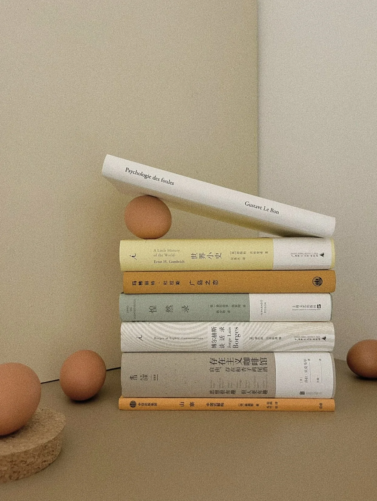 A stack of seven books arranged vertically on a surface, with a beige background. There are also two brown eggs and a small cork base nearby.