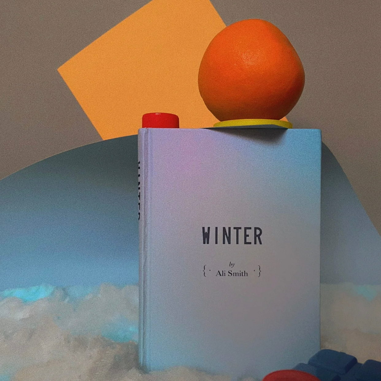 A book titled 'WINTER' by Ali Smith standing upright with an orange on top, a red bottle cap, and a yellow coaster on top of the book. Background includes colorful geometric shapes and clouds.