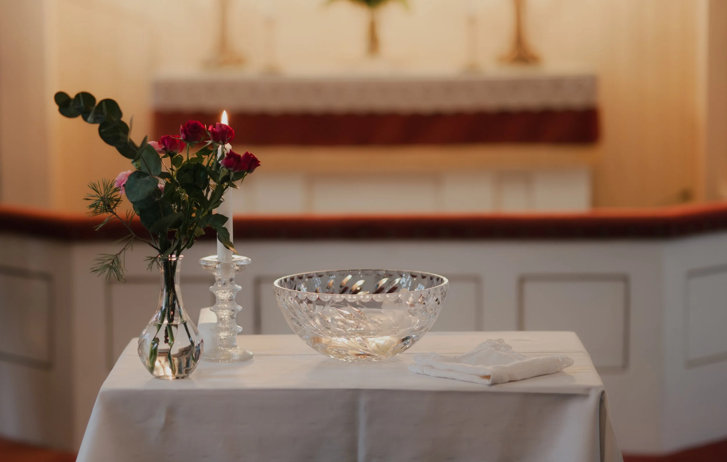 Kuva kastetilaisuudesta, jossa on kukkakimppu maljakossa, kynttilä ja kristalliallas pöydällä, kirkossa taustalla alttari ja kynttilöitä.
