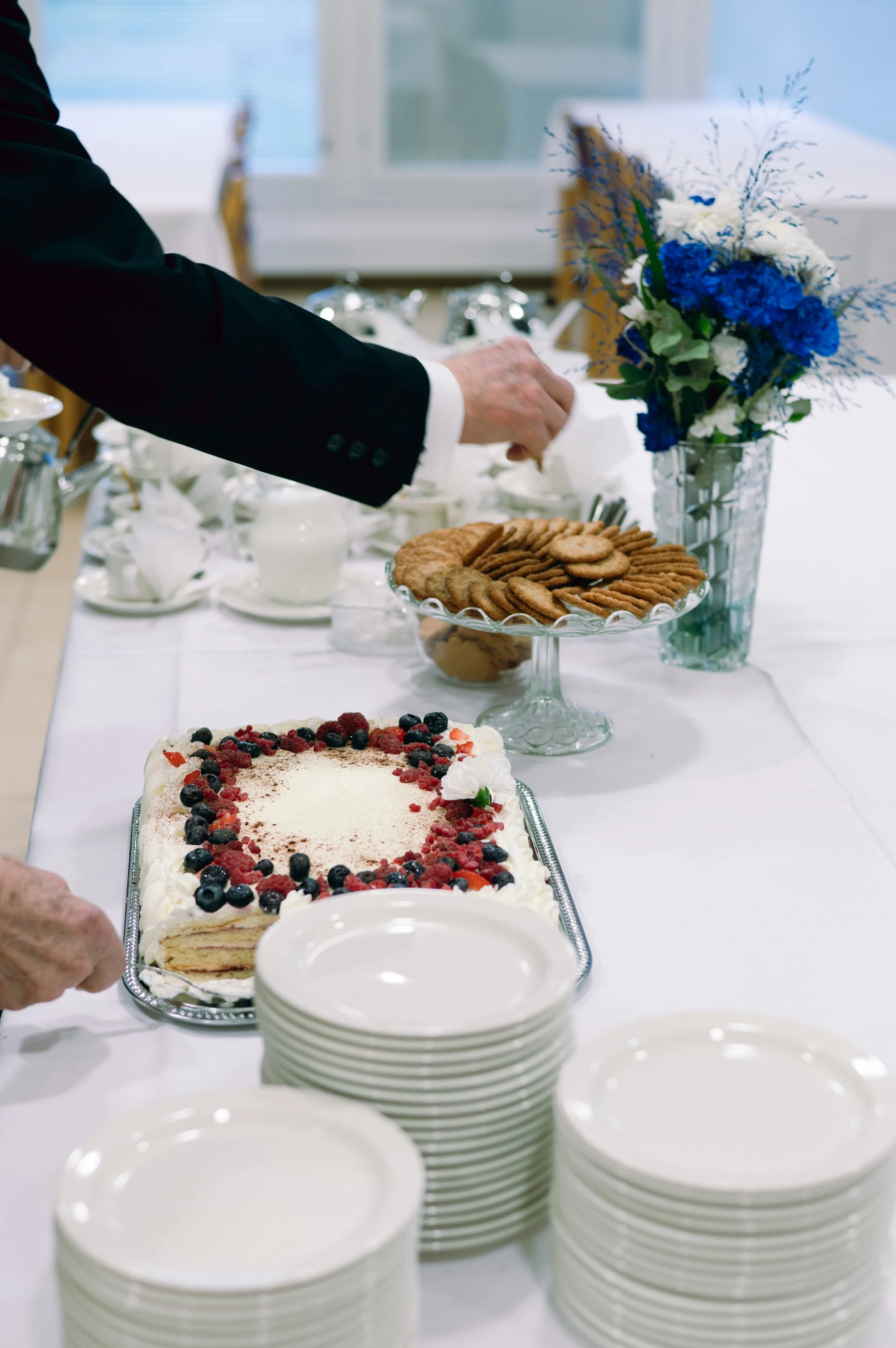 Henkilö ottaa keksirasiasta keksejä juhlapöydässä, jossa on myös kerroskakku ja kukkakimppu.