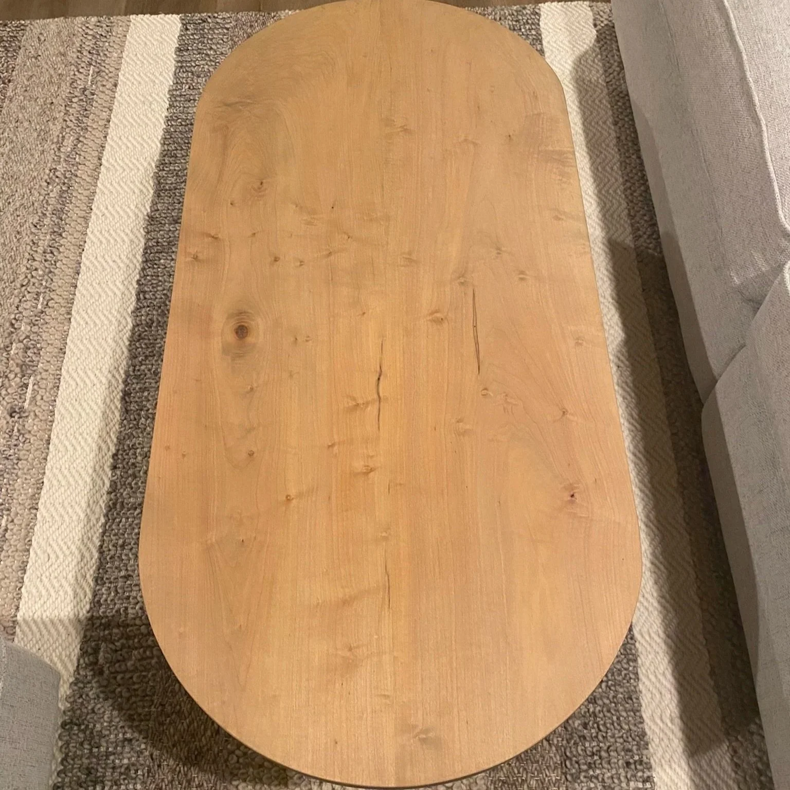 An oval wooden table with visible wood grain and knots, placed on a patterned area rug near a sofa.