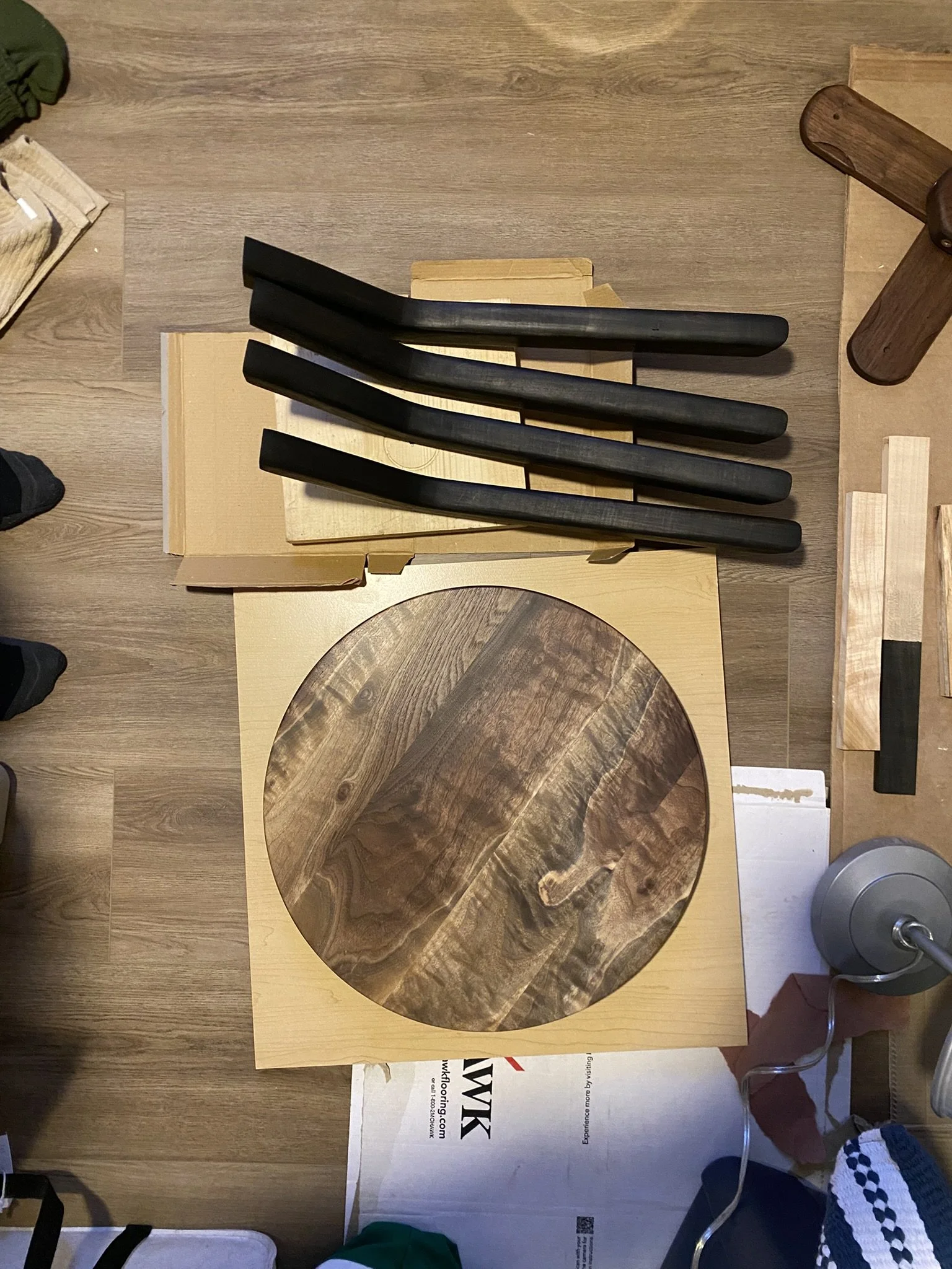 Top view of a wooden cutting board with a circular pattern, three black-handled knives, a wooden block, and various wooden tools and objects on a light wooden surface.