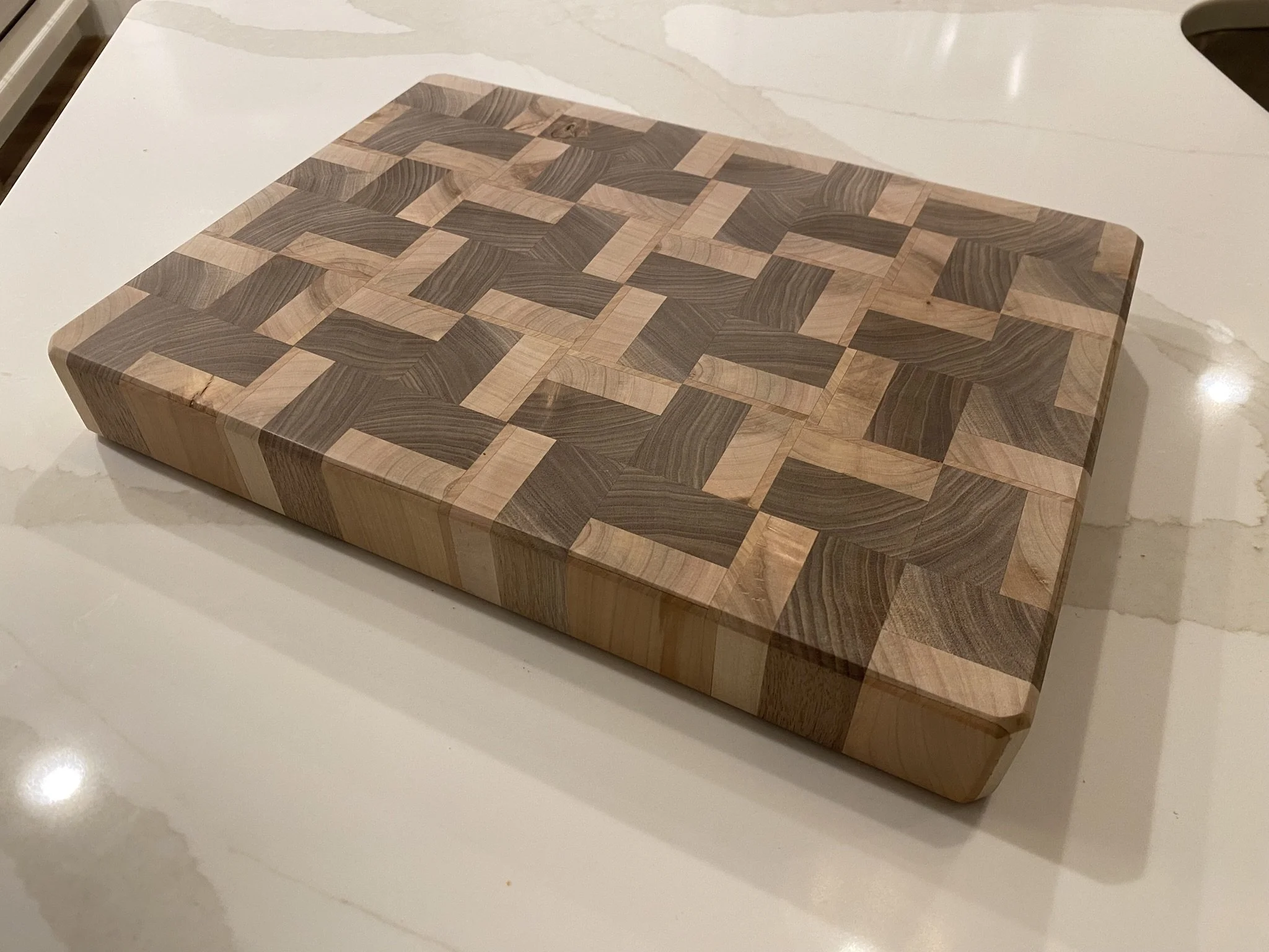 A rectangular wooden cutting board with a checkered pattern made of light and dark wood squares, placed on a marble countertop.