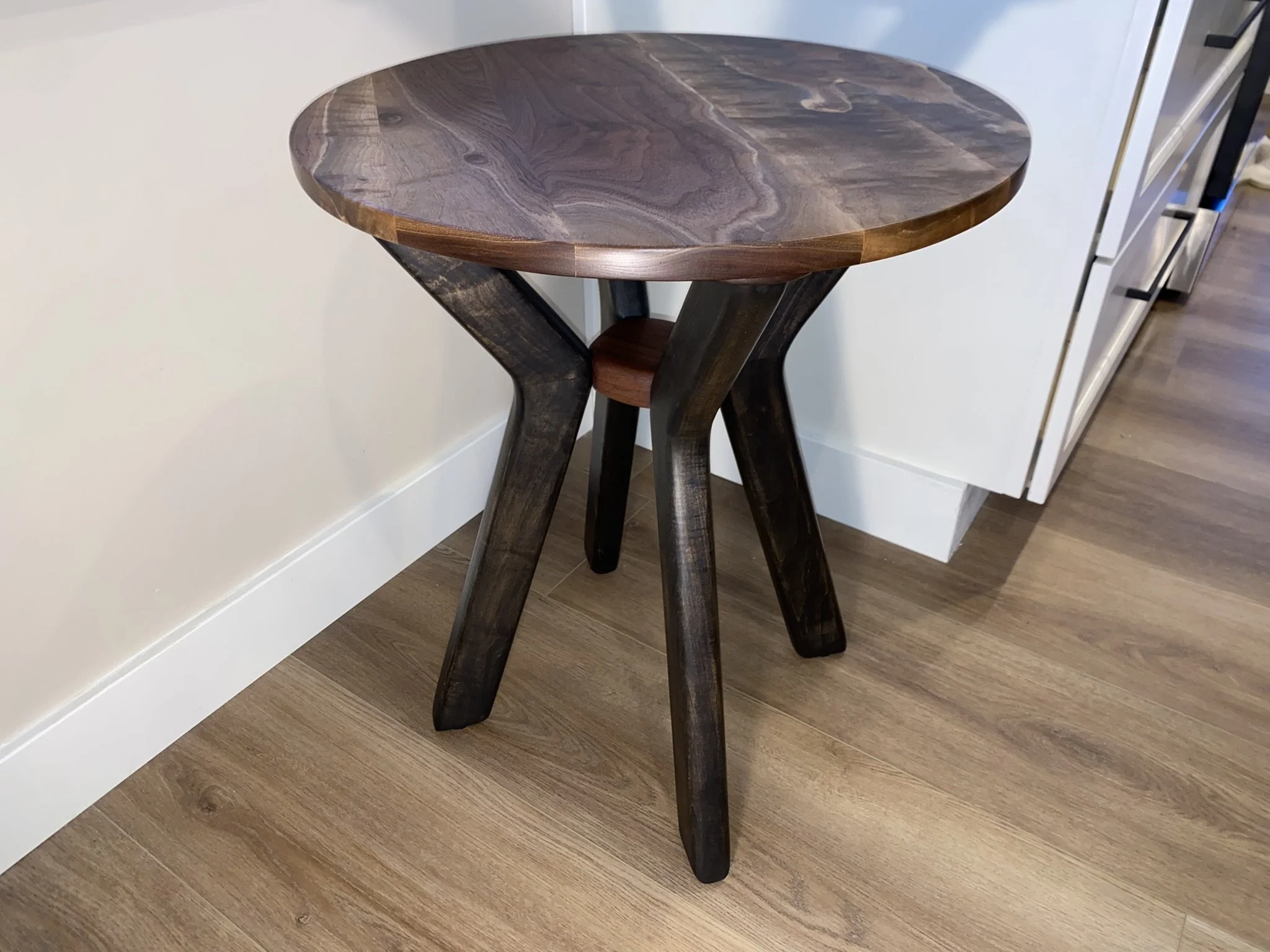 A small round wooden side table with three dark stained wooden legs, placed on a wooden floor next to a white wall and cabinet.