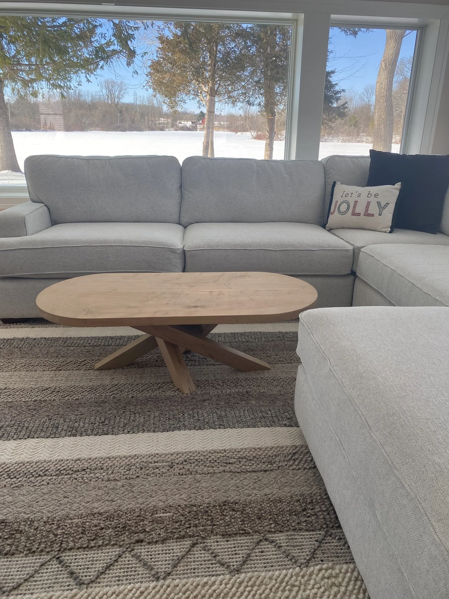 A living room with a light gray sectional sofa, a wooden coffee table, and large windows showing a snowy outdoor scene with trees and a frozen landscape.