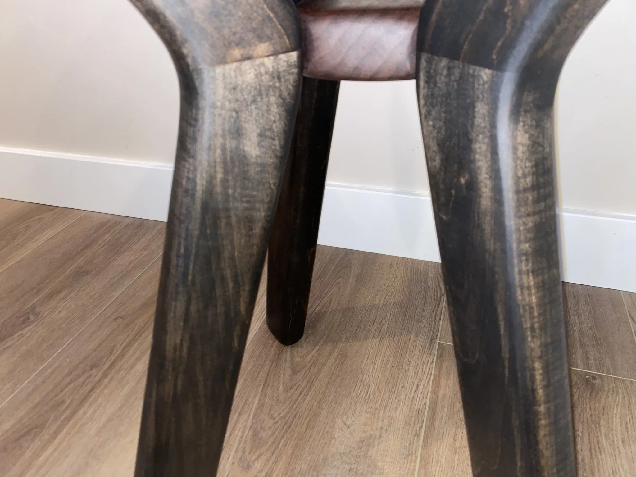 Close-up of the legs of a wooden chair with a dark finish on a wooden floor, against a white wall with baseboard.