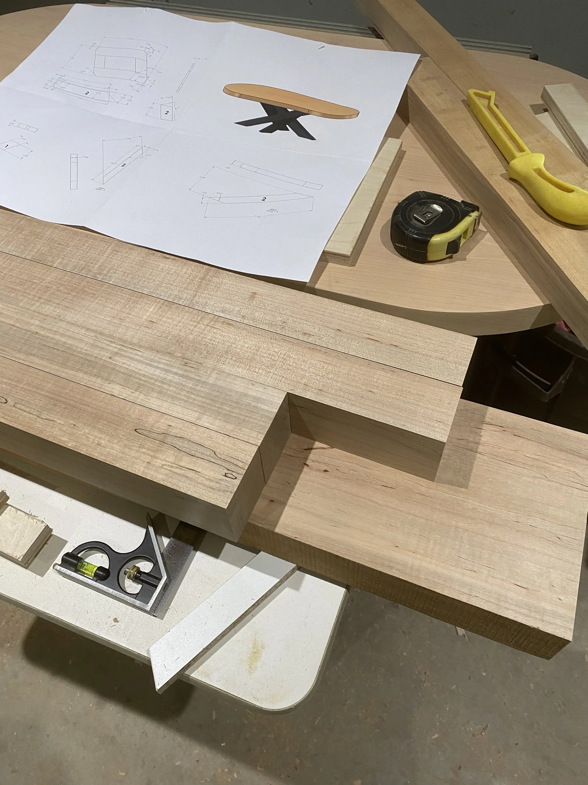 Carpentry workspace with wood pieces, a sheet of paper with a design drawing, a yellow hand saw, a tape measure, and a square tool.