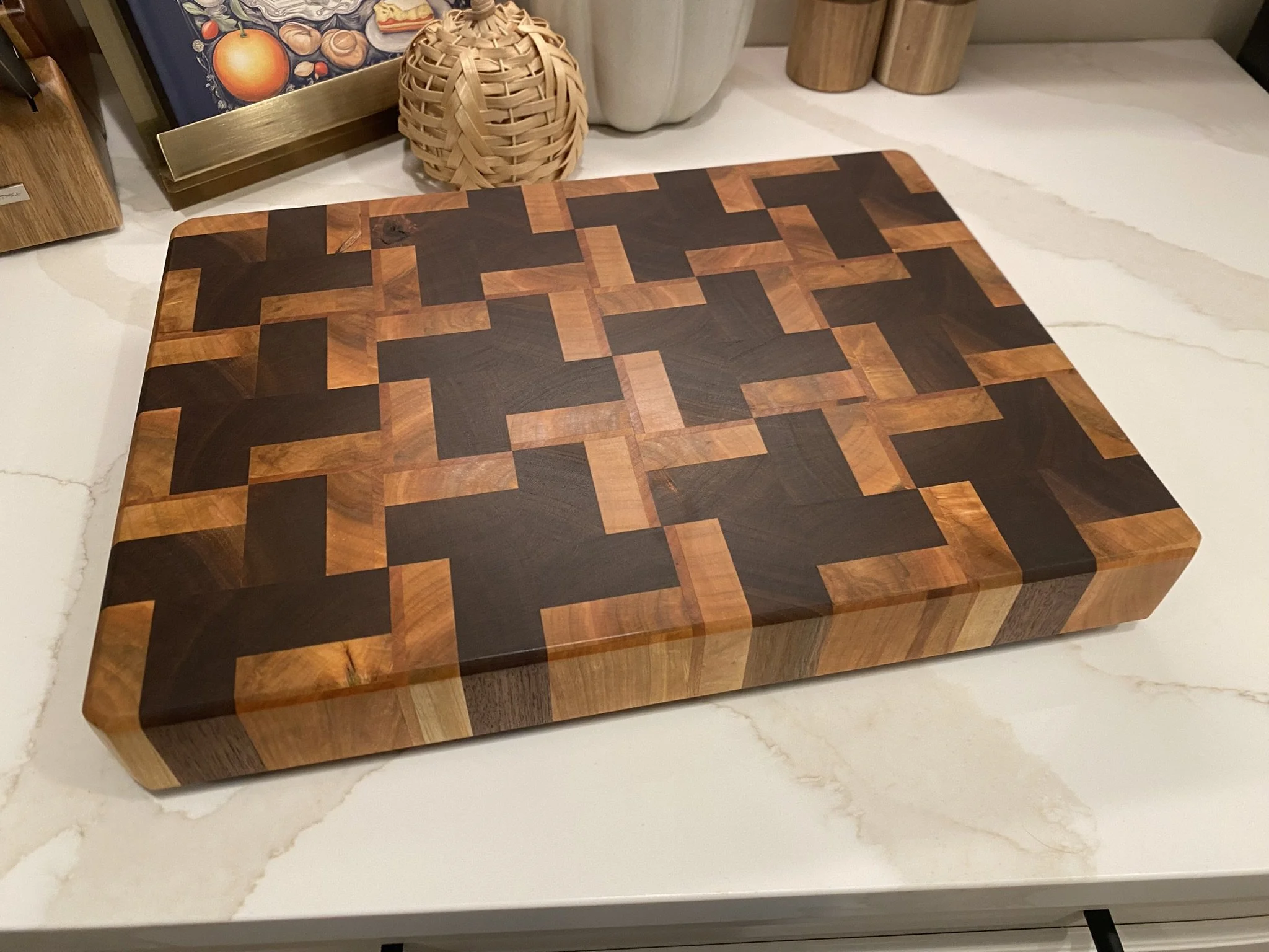 A wooden cutting board with a geometric pattern of alternating dark and light wood in a herringbone style, placed on a white marble countertop.