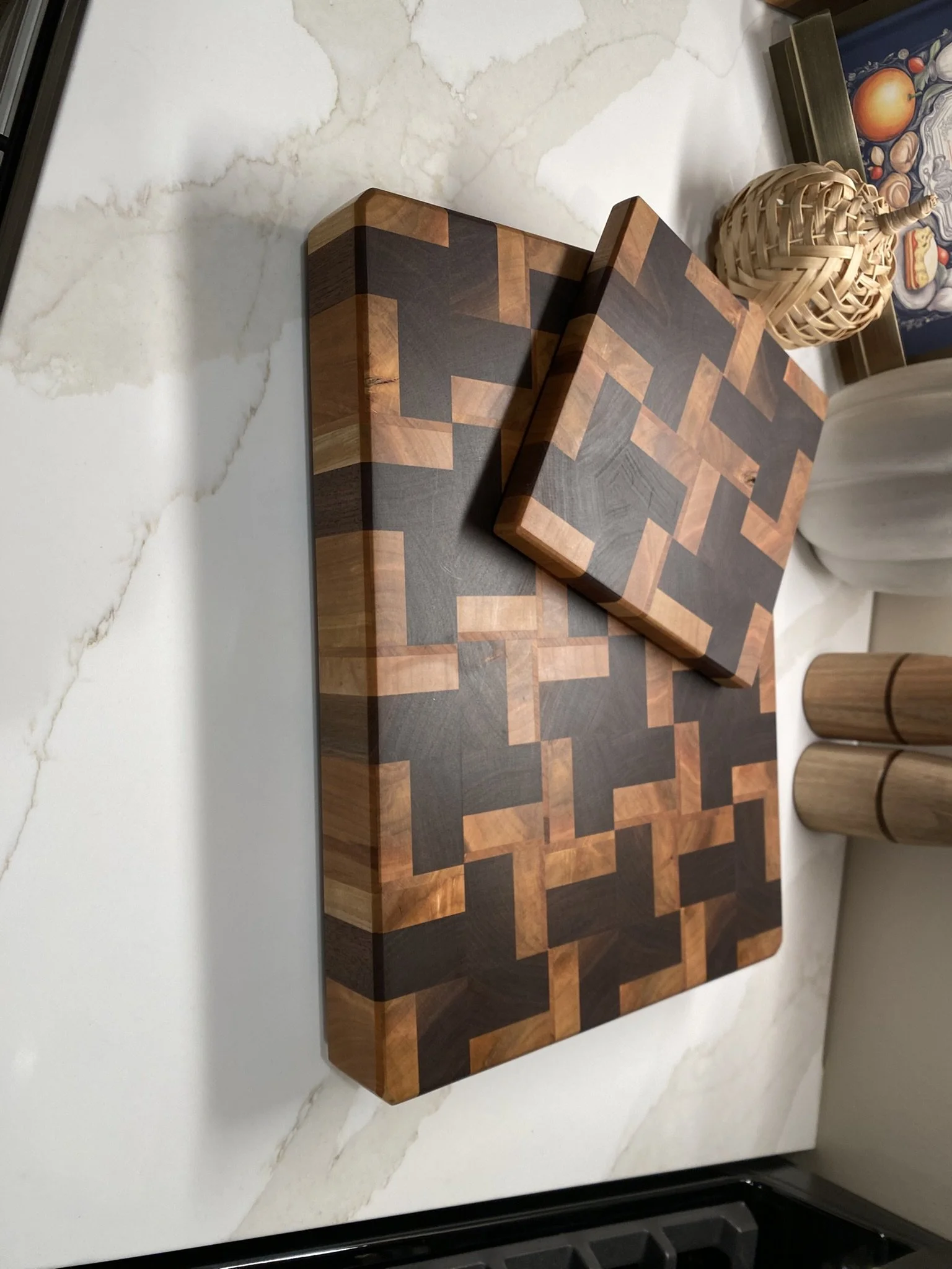 A geometric wood cutting board with a matching smaller piece placed over it, both featuring an intricate pattern of alternating light and dark wood squares, on a white marble surface with decorative vases nearby.