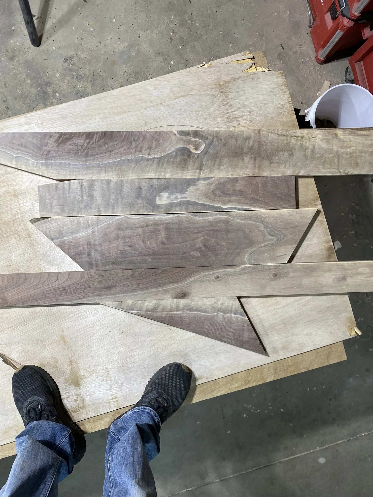 A top-down view of a workbench with several pieces of wood, some partially cut, and a person's feet in black sneakers and jeans, in a workshop.