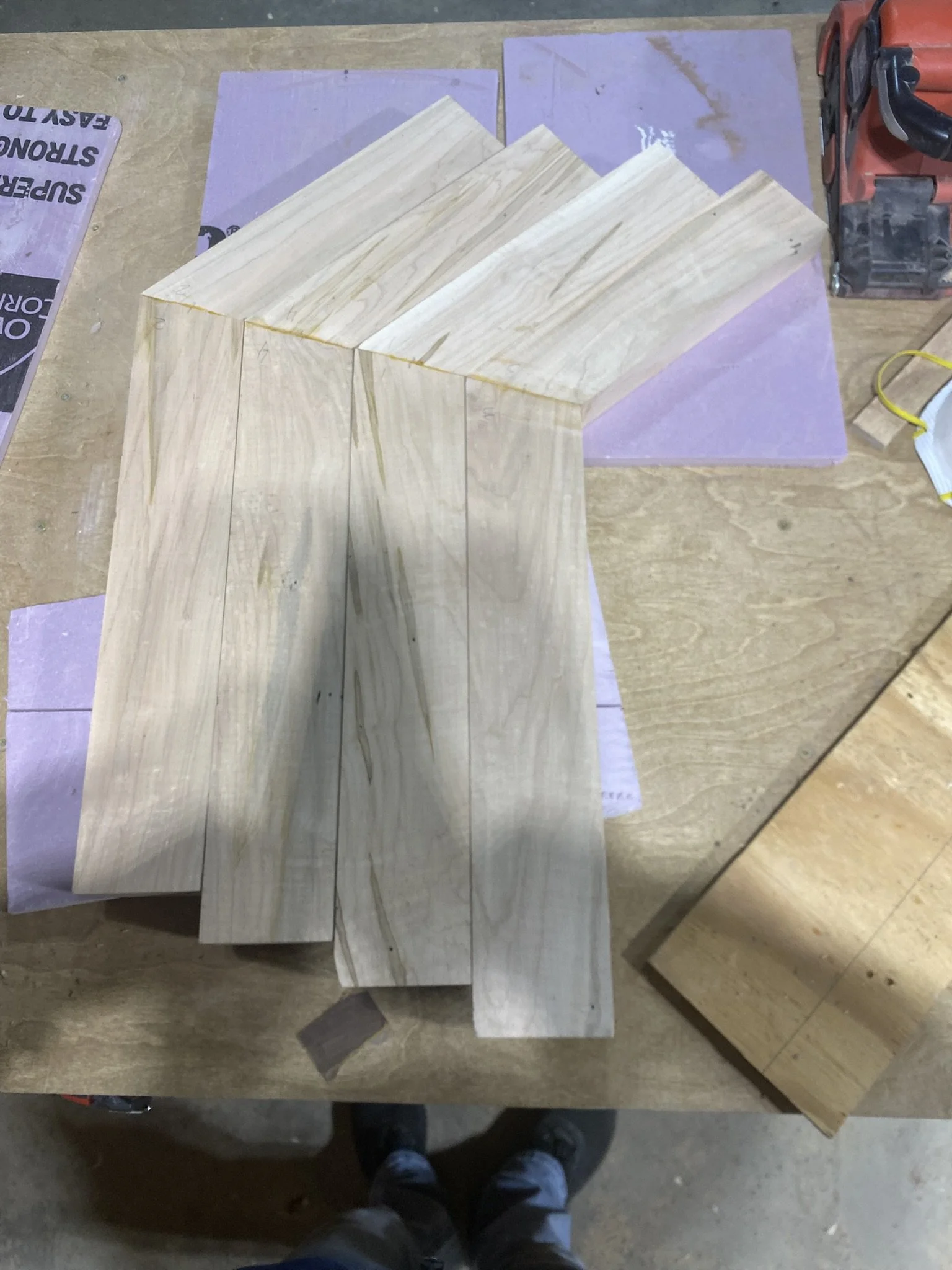 Several light-colored wooden boards on a workbench, some pink and purple sanding papers, a red power saw, and a face mask.