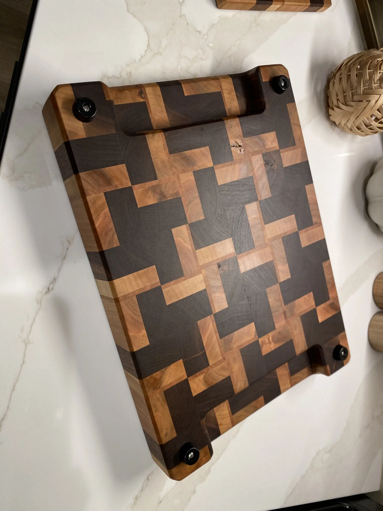 A rectangular wooden cutting board with a geometric pattern of light and dark wood pieces, placed on a white marble countertop.