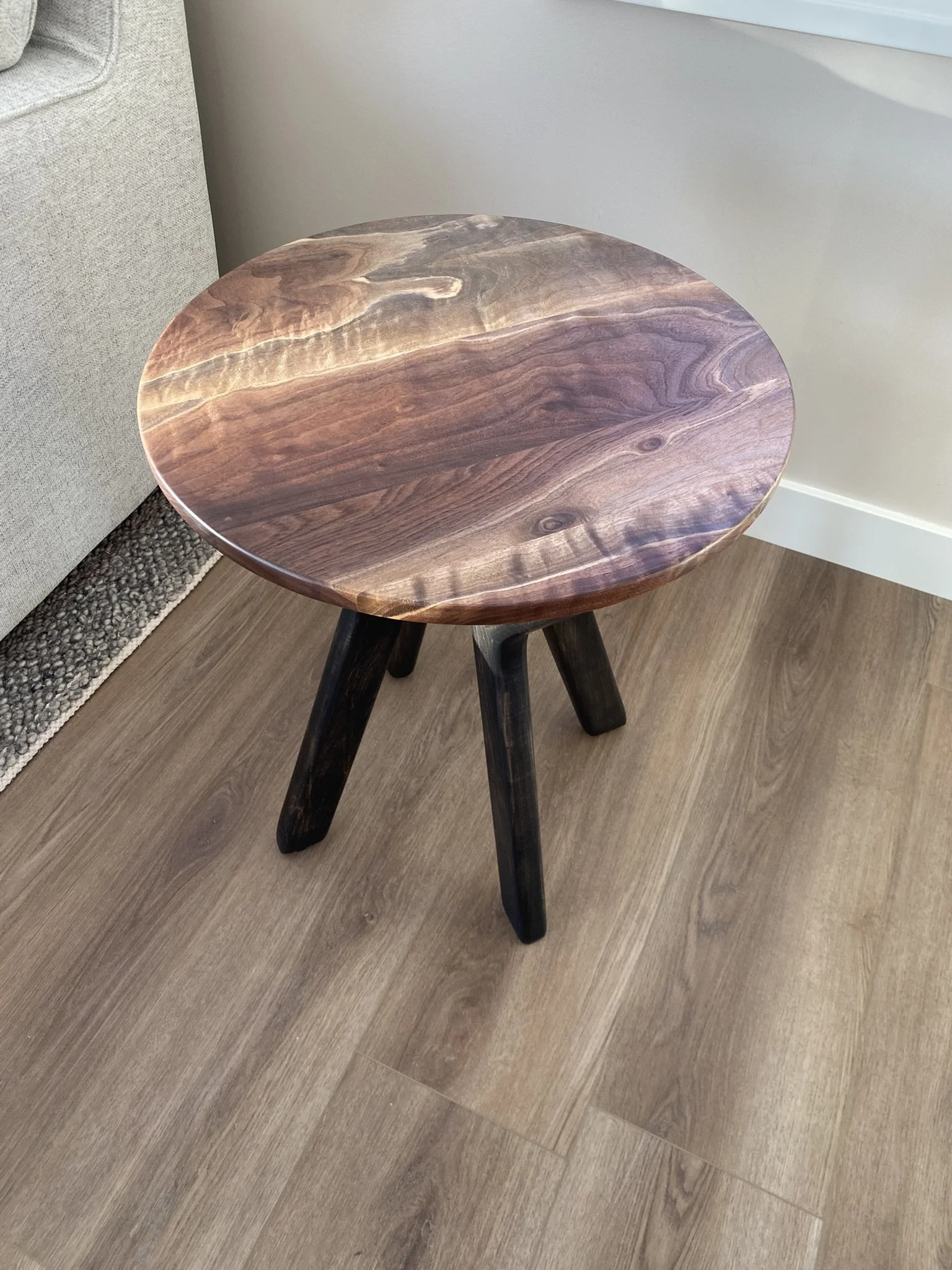 A small round wooden side table with a dark wood finish, placed on a light wooden floor next to a light-colored sofa.