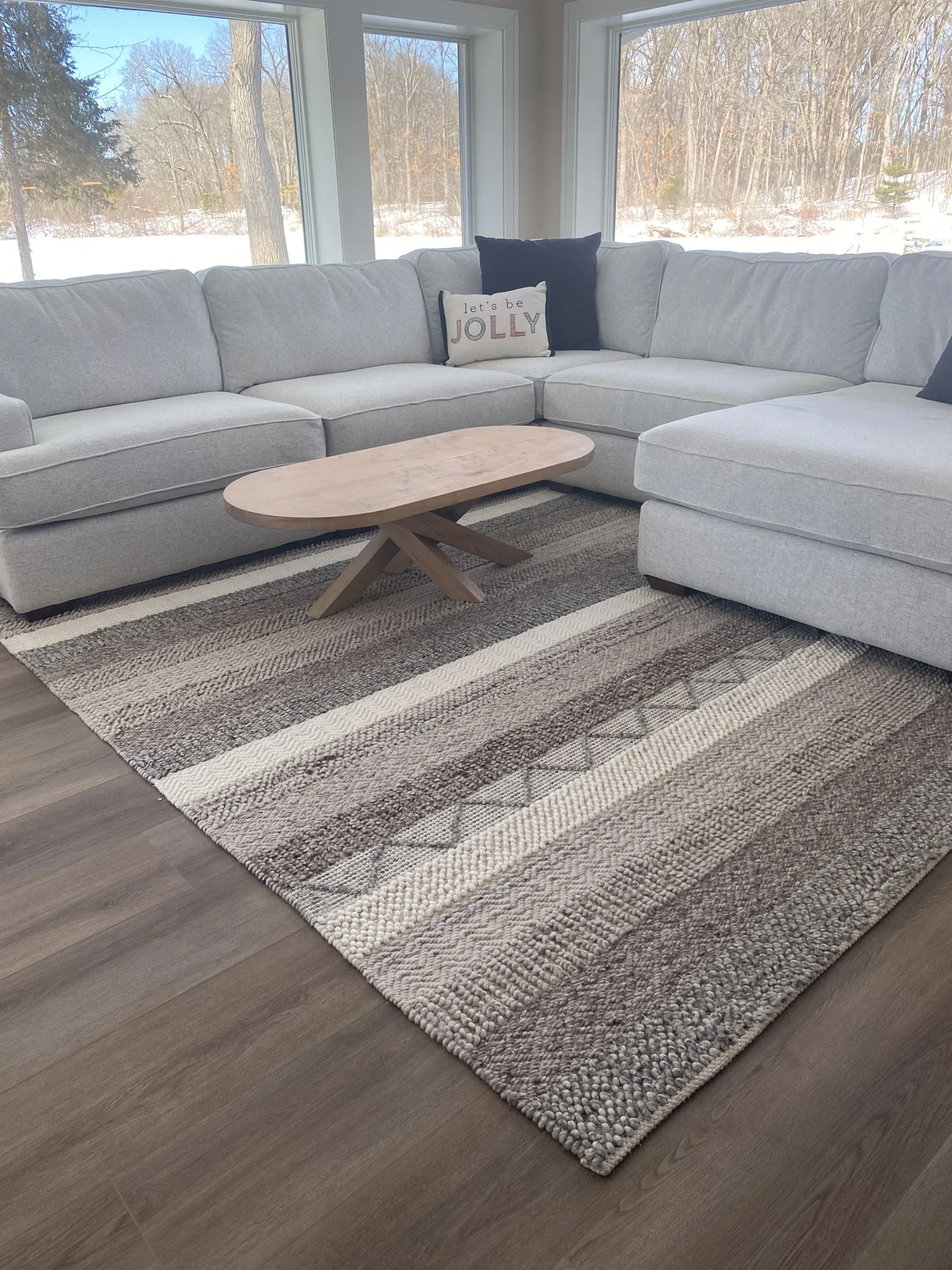Living room with a large light gray sectional sofa, a wooden coffee table, and a patterned area rug. Large windows offer a view of a snowy outdoor landscape with trees.