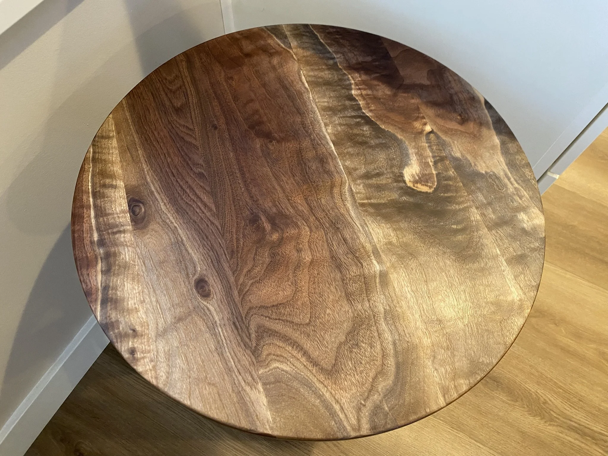 Close-up of a round wooden table with a natural grain pattern, placed next to a light gray wall and a white piece of furniture.