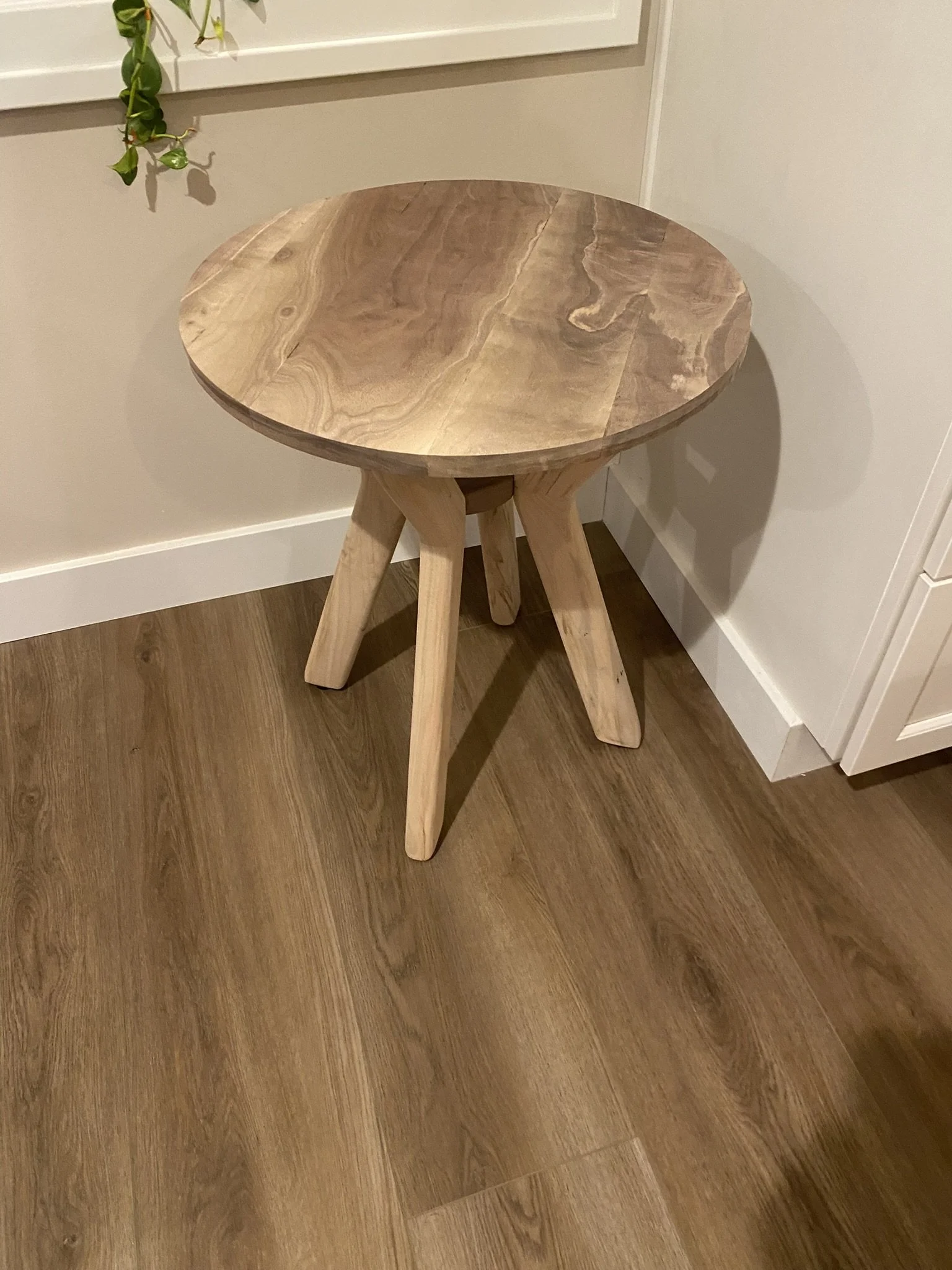 A small round wooden table with three angled legs, placed near a wall and a window with some green plant leaves hanging.