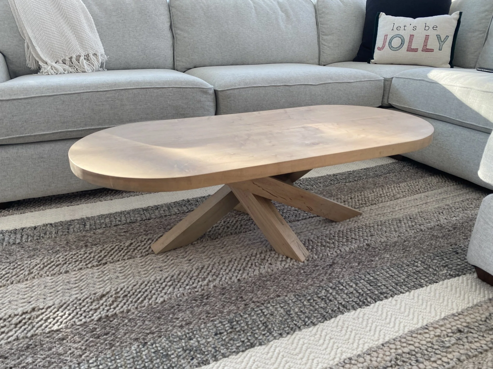 Wooden coffee table on a textured rug in front of a light gray sofa with pillows, including one with the words 'let's be JOLLY'.