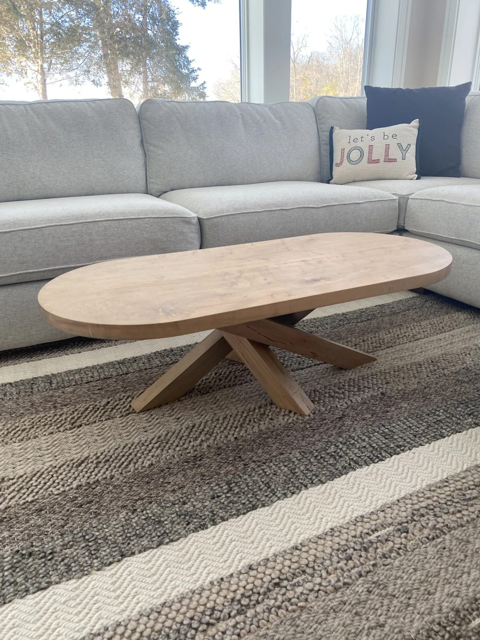 Living room with a light gray sectional sofa, a wooden oval coffee table, a black pillow, and a decorative pillow with the words "let's be JOLLY." The room has a large window with trees and a blue sky in the background.