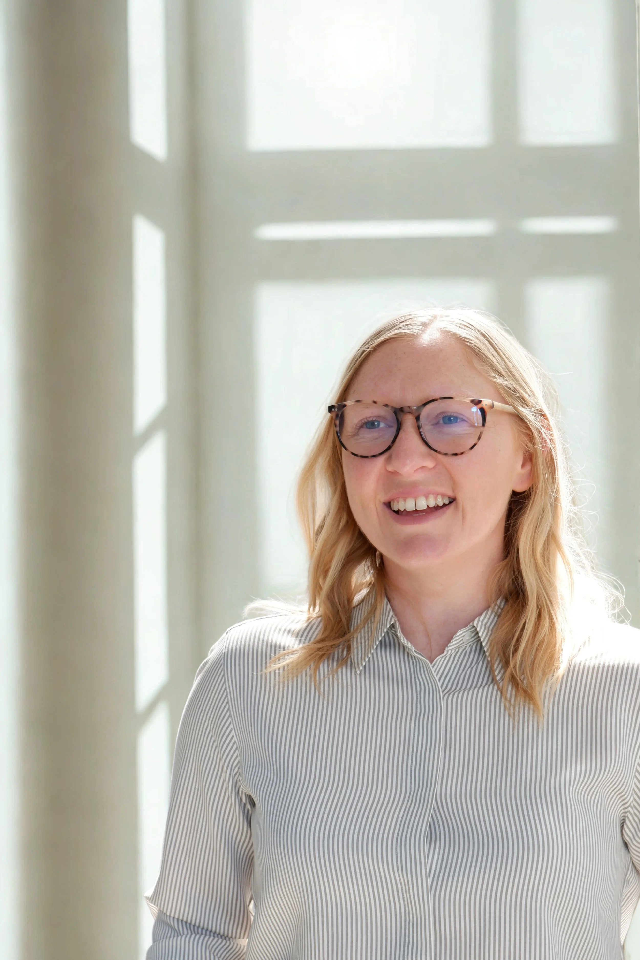 A woman with blonde hair and glasses, wearing a striped shirt, smiling and looking to her right in front of a window with natural light.