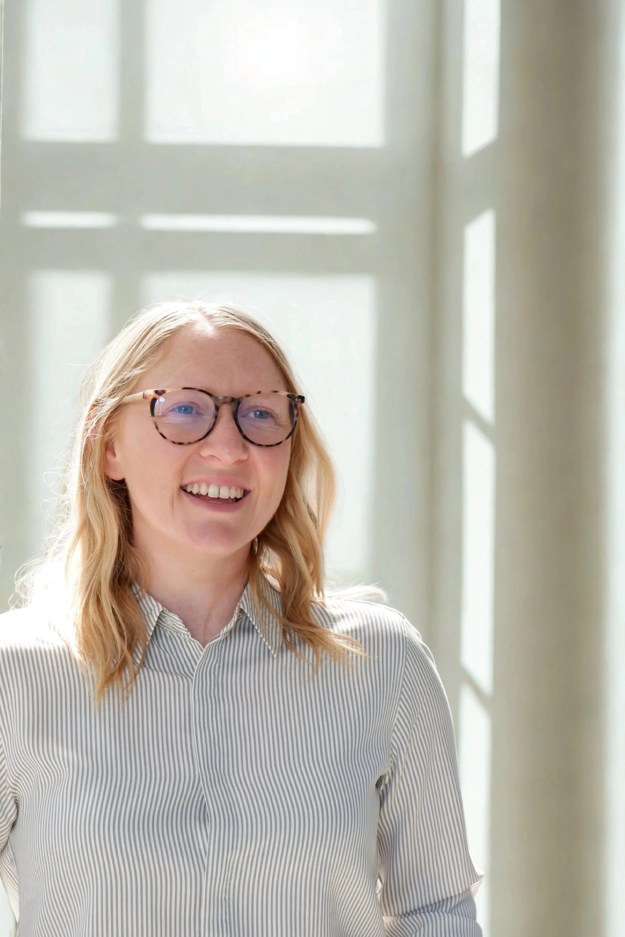 A woman with blonde hair, wearing glasses and a striped shirt, smiling indoors with sunlight coming through a window behind her.