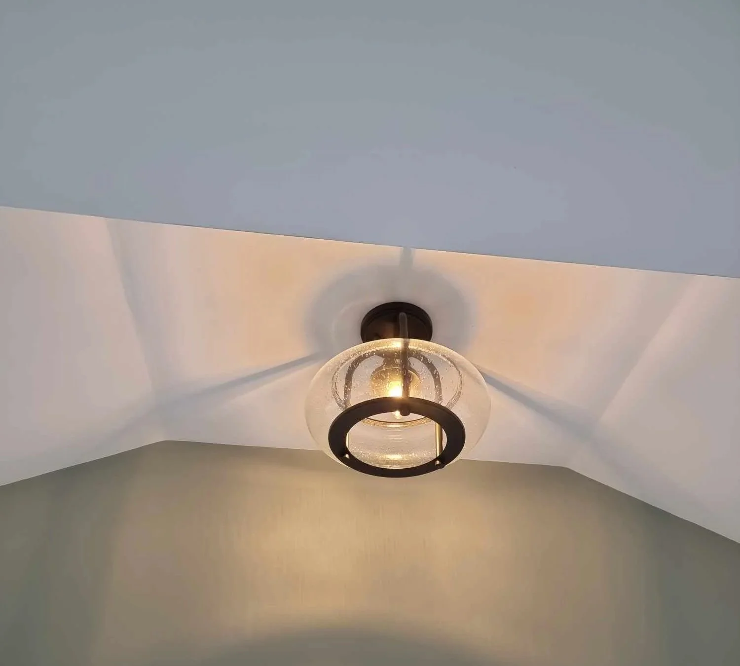 Ceiling light fixture with a round glass shade and a black circular frame, mounted on a white ceiling with  a vaulted design, illuminated and casting shadows.